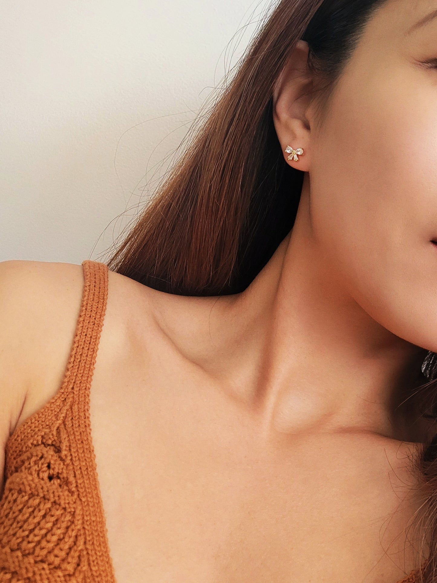 Close up of a woman's ear wearing dainty bow stud earrings with sparkling cubic zirconia and gold plating.
