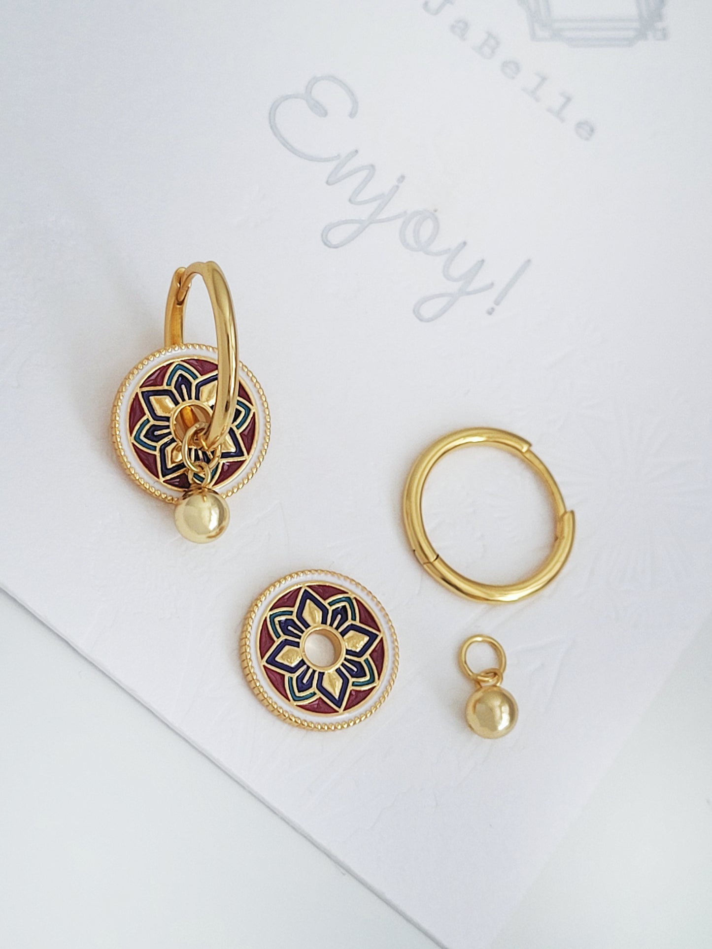 Gold hoop earrings featuring a tile pattern disc and a gold ball dangle, displayed on a white surface showcasing their elegant and unique artisan design.
