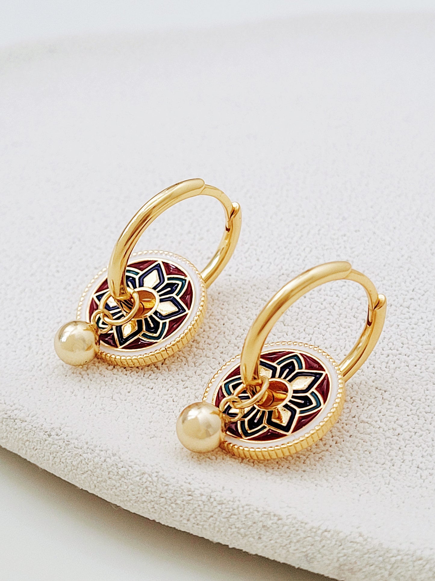 Gold hoop earrings featuring a tile pattern disc and a gold ball dangle, displayed on a white surface showcasing their elegant and unique artisan design.