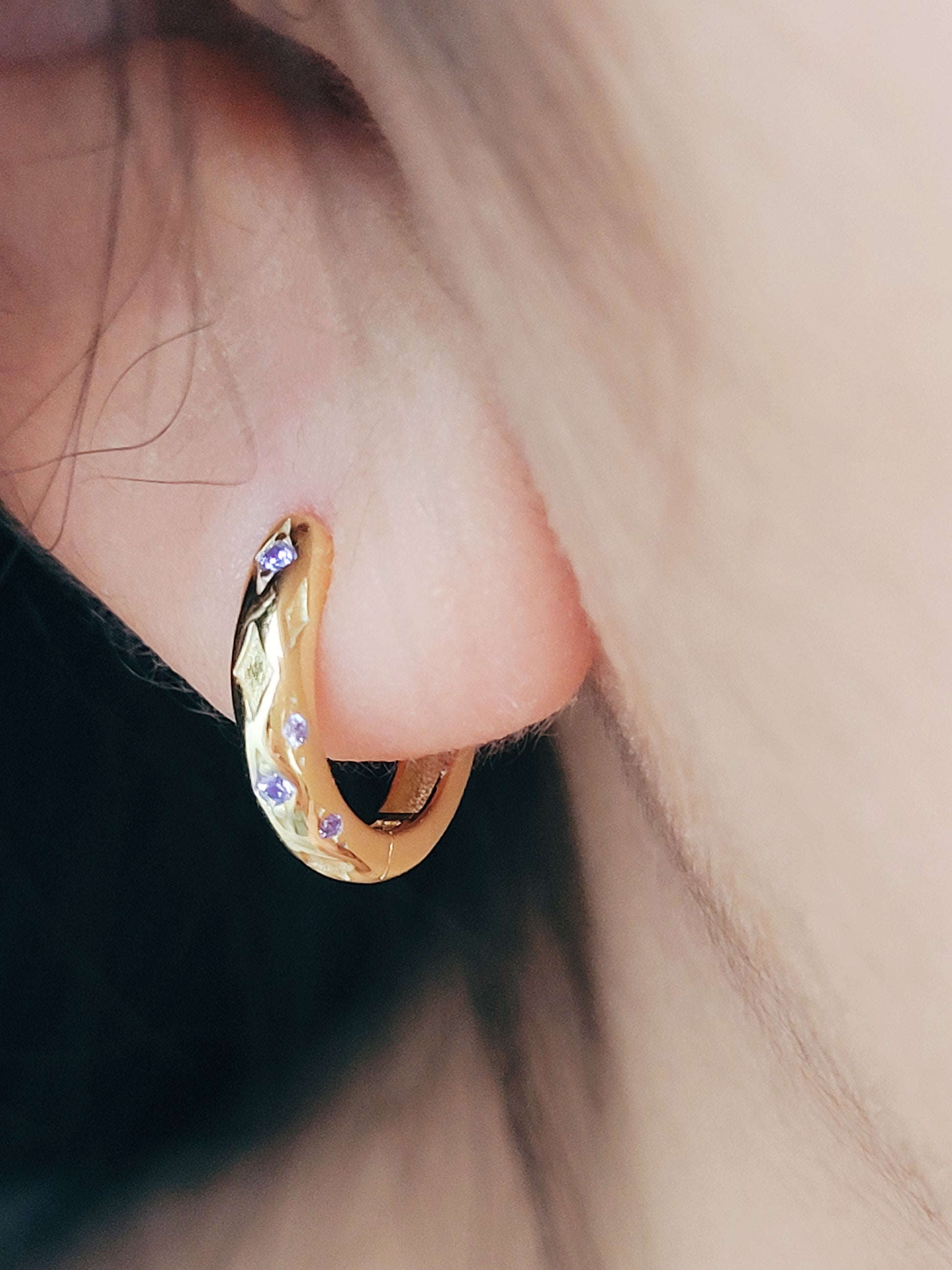 Close up of a woman wearing 18k gold plated sterling silver huggie hoop earrings with purple crystals, featuring a cosmic star inspired design.