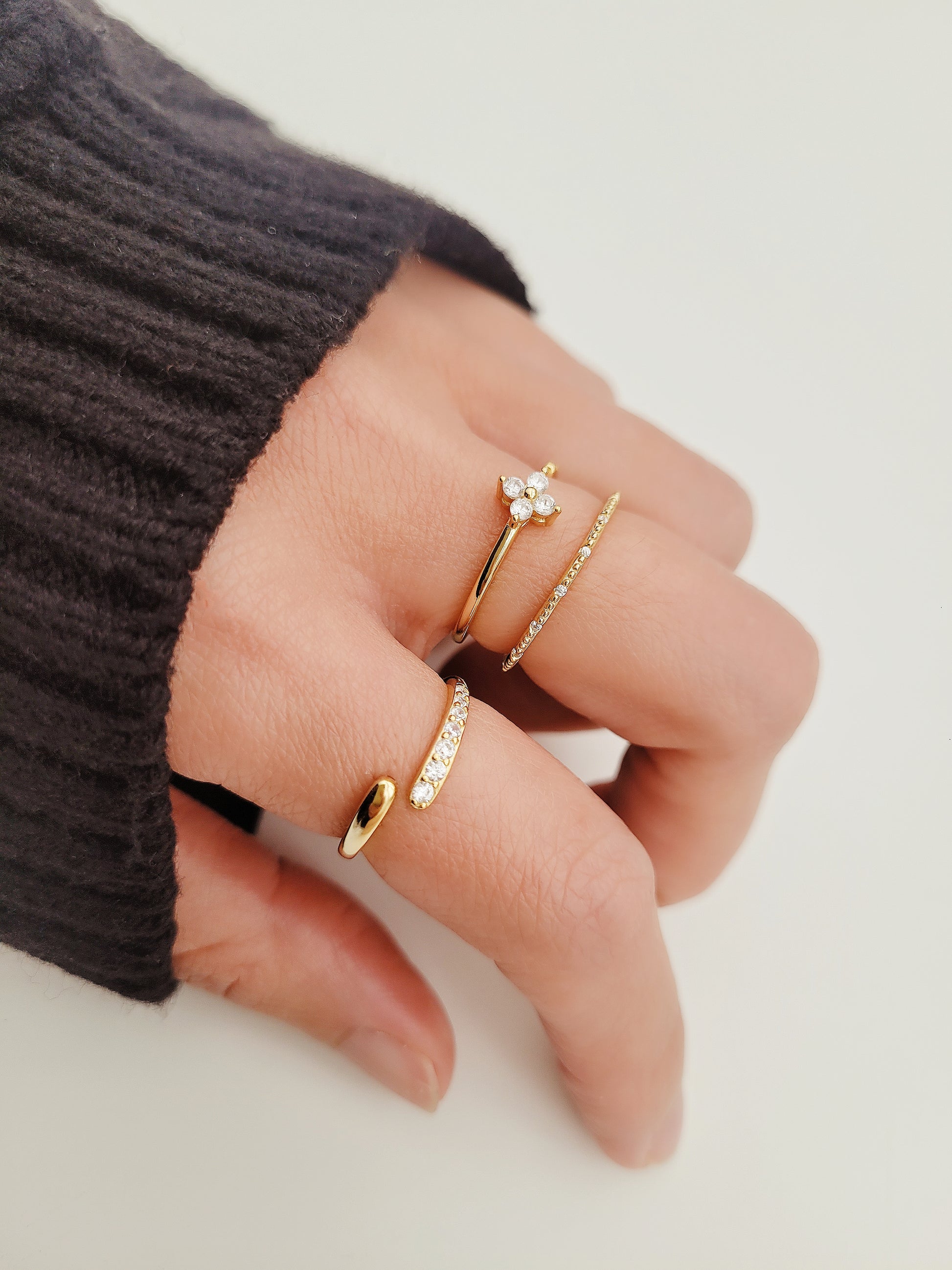 A hand wearing three elegant stacking rings, each featuring gold accents. One ring is open-ended with a sleek, minimalistic design, another is encrusted with small sparkling stones, and the third ring has a delicate clover-shaped cluster of gemstones.
