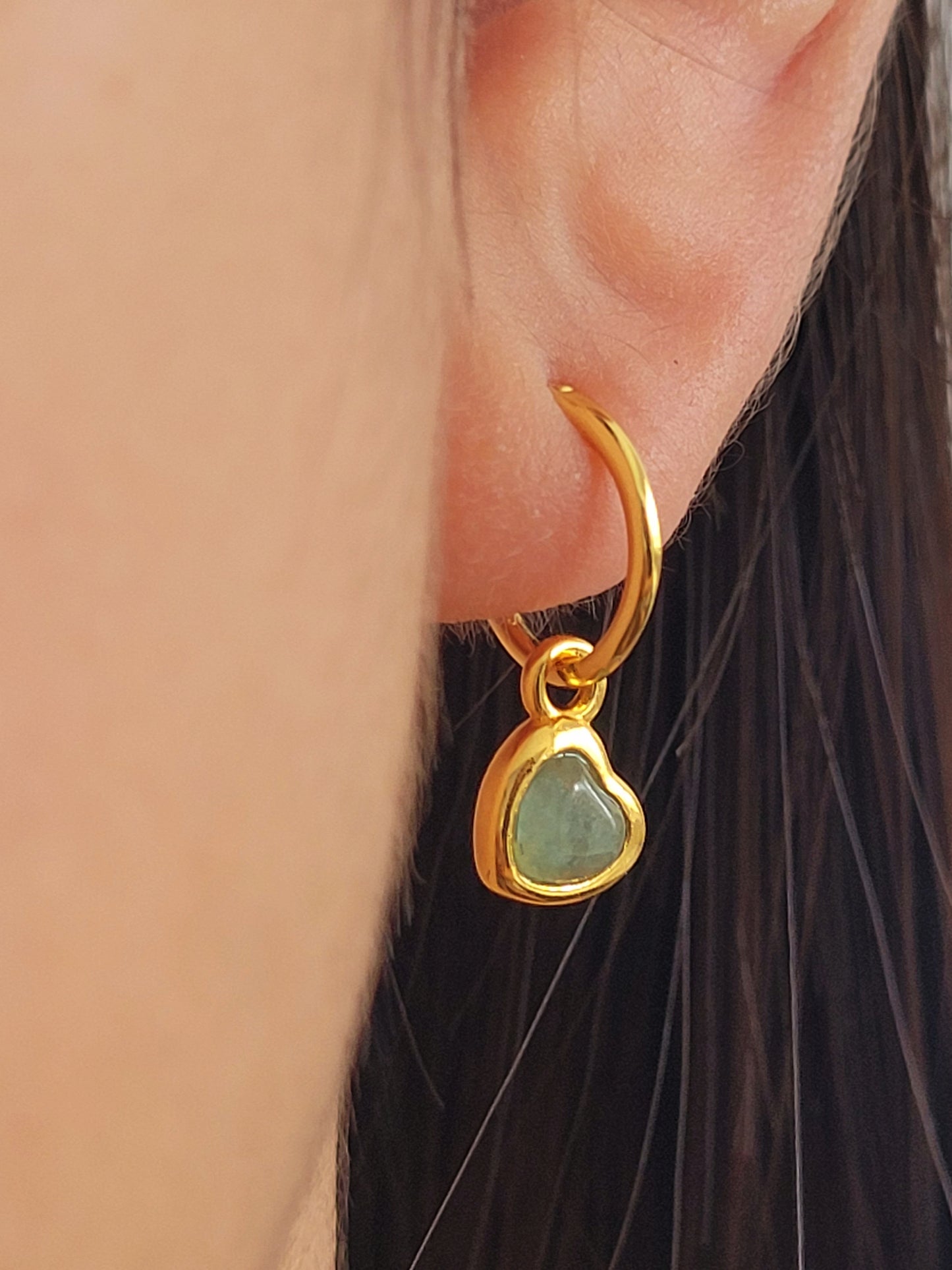 Gold heart earring with a green gemstone worn by an ear