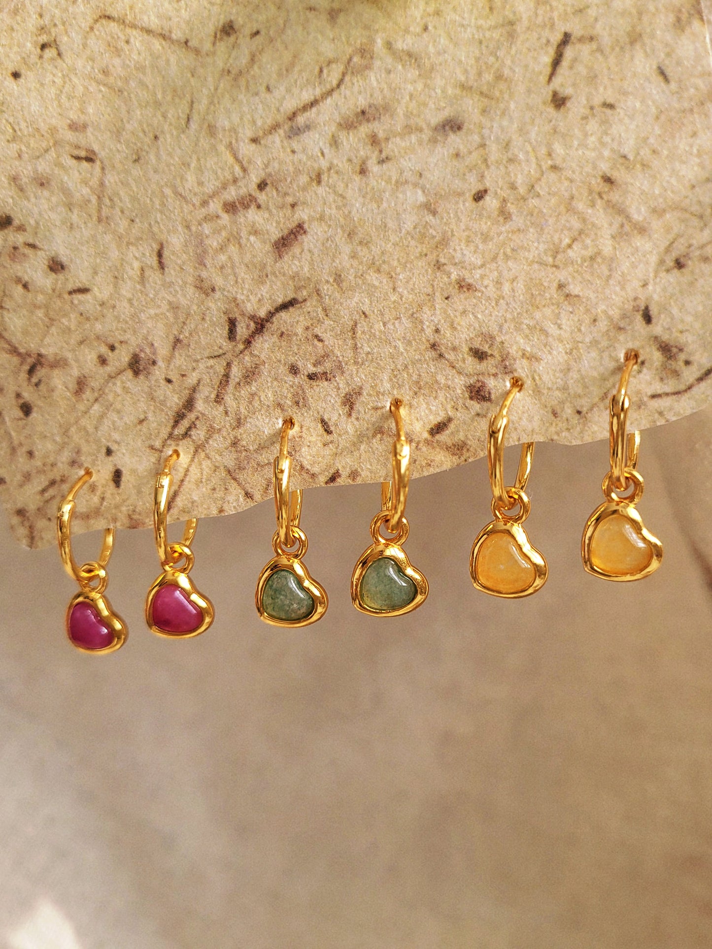Set of gold heart earrings with gemstone drops on a textured beige background