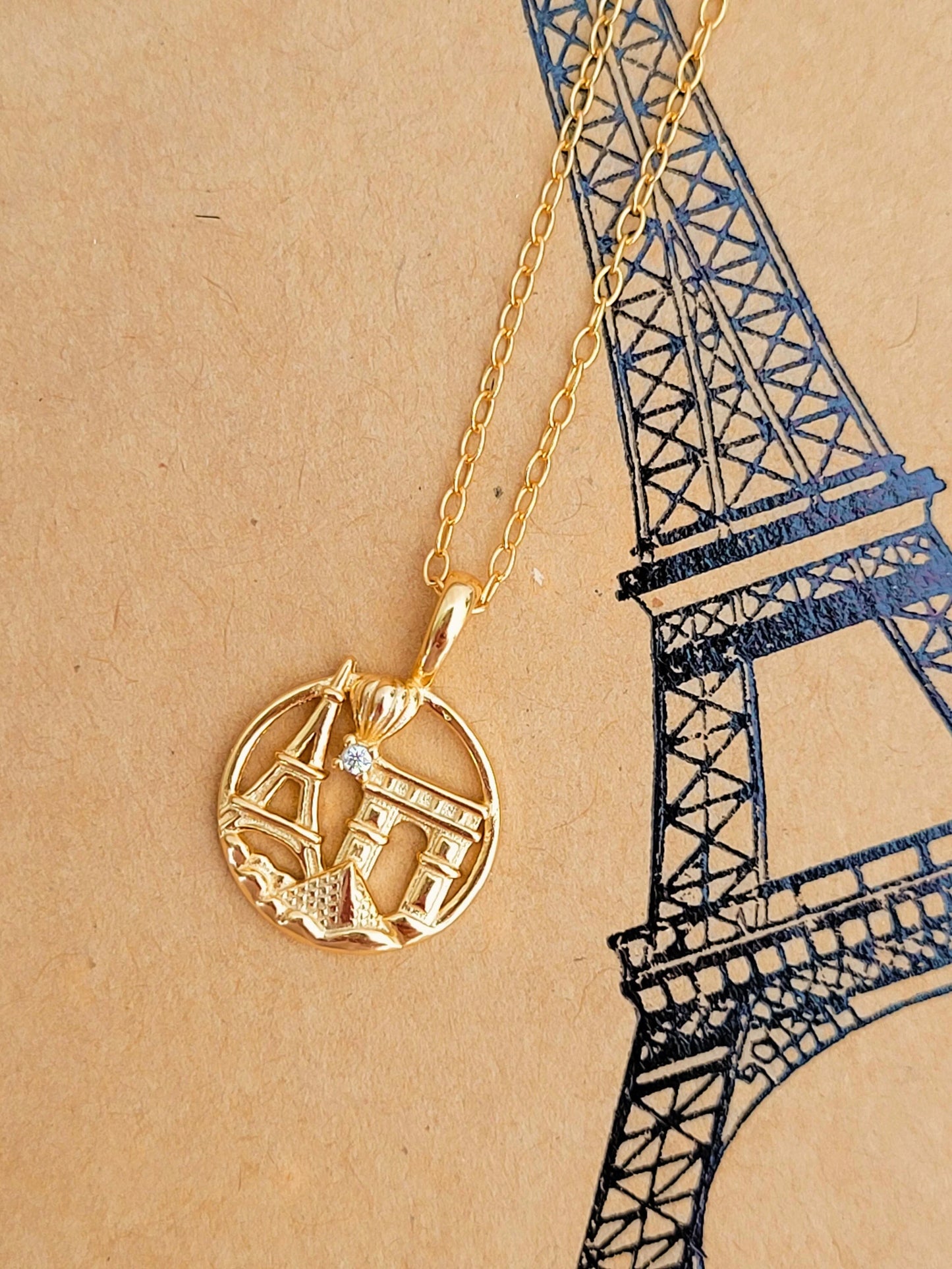 Gold necklace with a Paris-themed pendant on a beige background with a black outline of the Eiffel Tower.