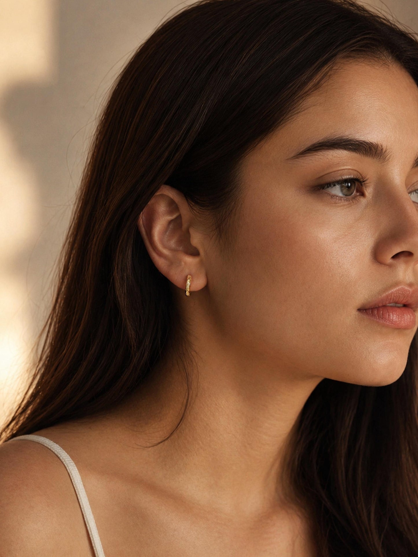 A close-up side profile of a woman with long dark brown hair wearing a tiny gold hoop earring, modern and stylish jewellery, set against a warm beige background with soft shadows.