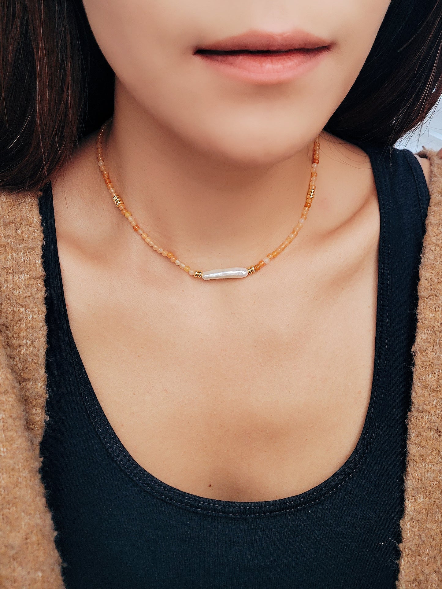 Close-up of a woman wearing an orange agate beaded necklace with a central baroque pearl. The necklace features small, round orange beads accented by gold elements and a single elongated, irregular-shaped white pearl in the center.