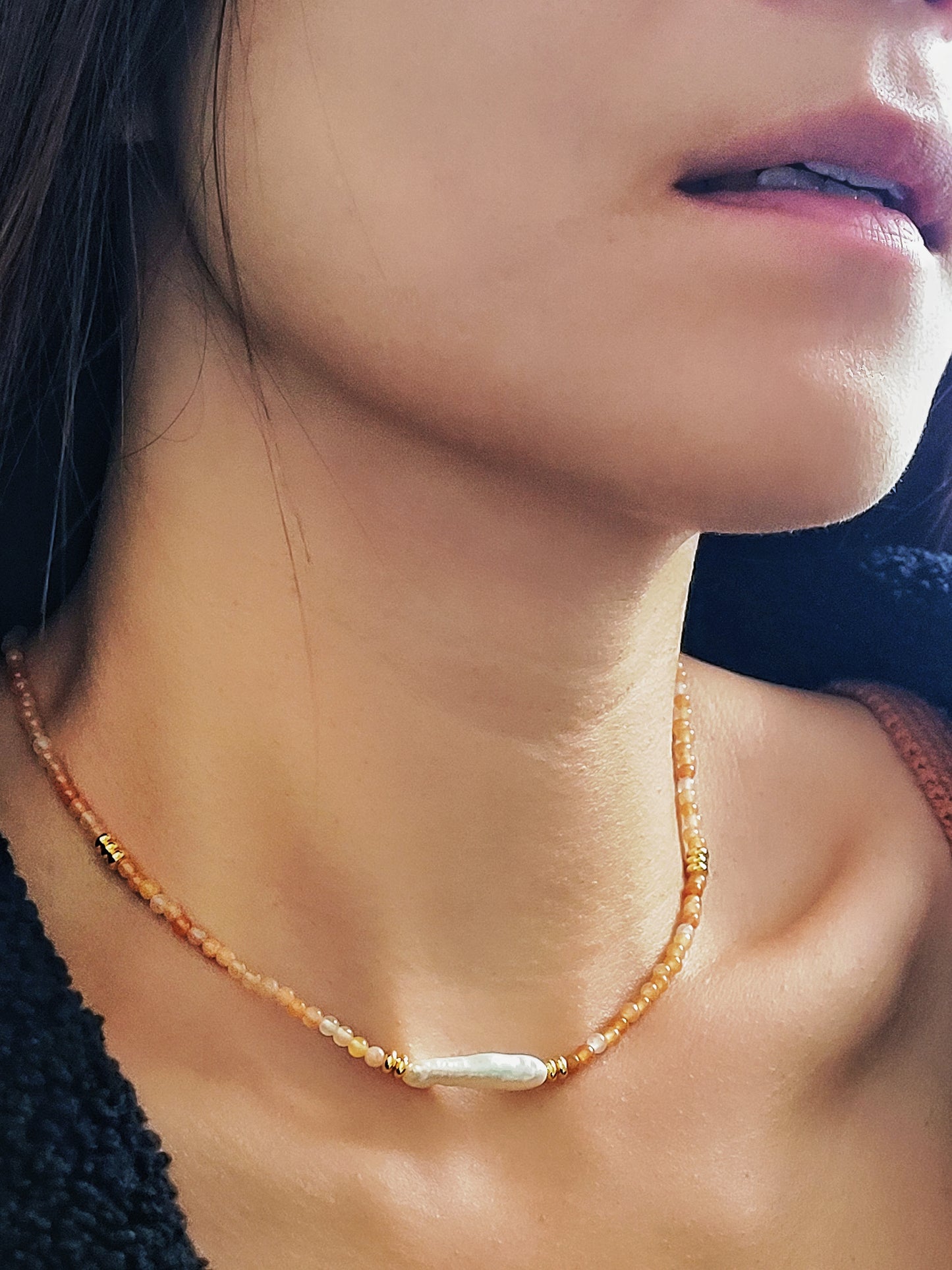 Close-up of a woman wearing an orange agate beaded necklace with a central baroque pearl. The necklace features small, round orange beads accented by gold elements and a single elongated, irregular-shaped white pearl in the center.