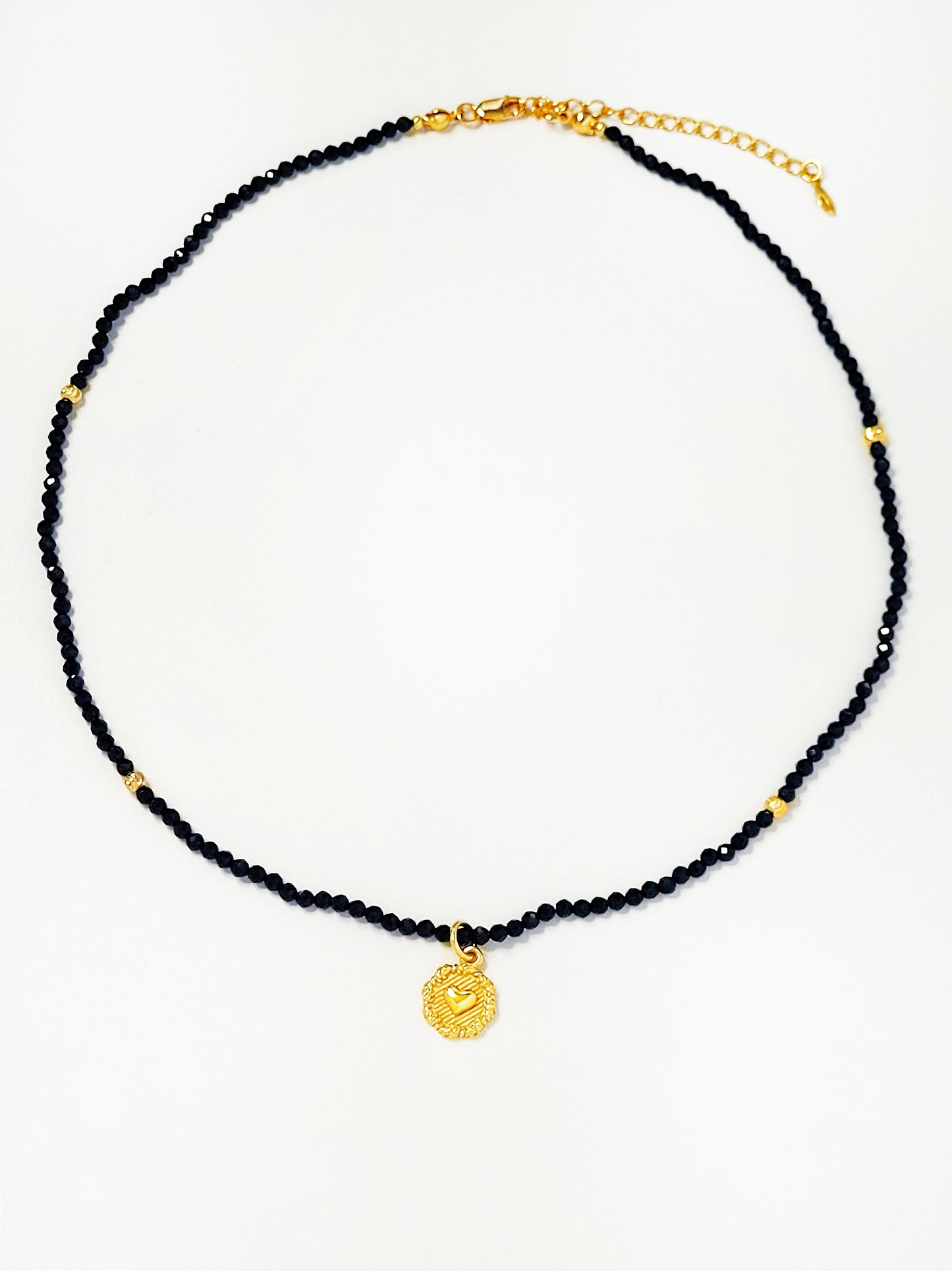 Close-up of a black spinel beaded choker with a gold puff heart pendant, featuring extra shine black beads and intricate gold detailing. The necklace lies on a white background, showcasing its elegance and refined craftsmanship.