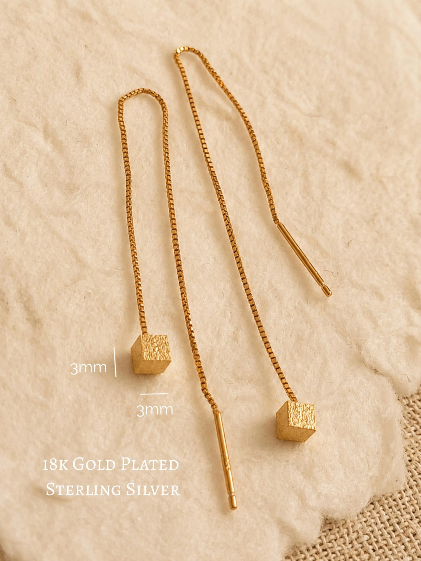 Close-up of two gold box chain threader earrings showing the fine detail of the textured, glittering square cube charms suspended at the ends against a warm, neutral background.