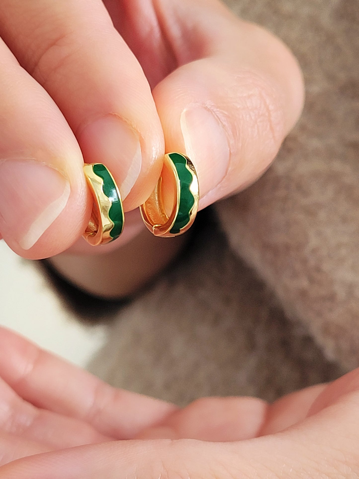Gold and green earrings held between fingers against a neutral background
