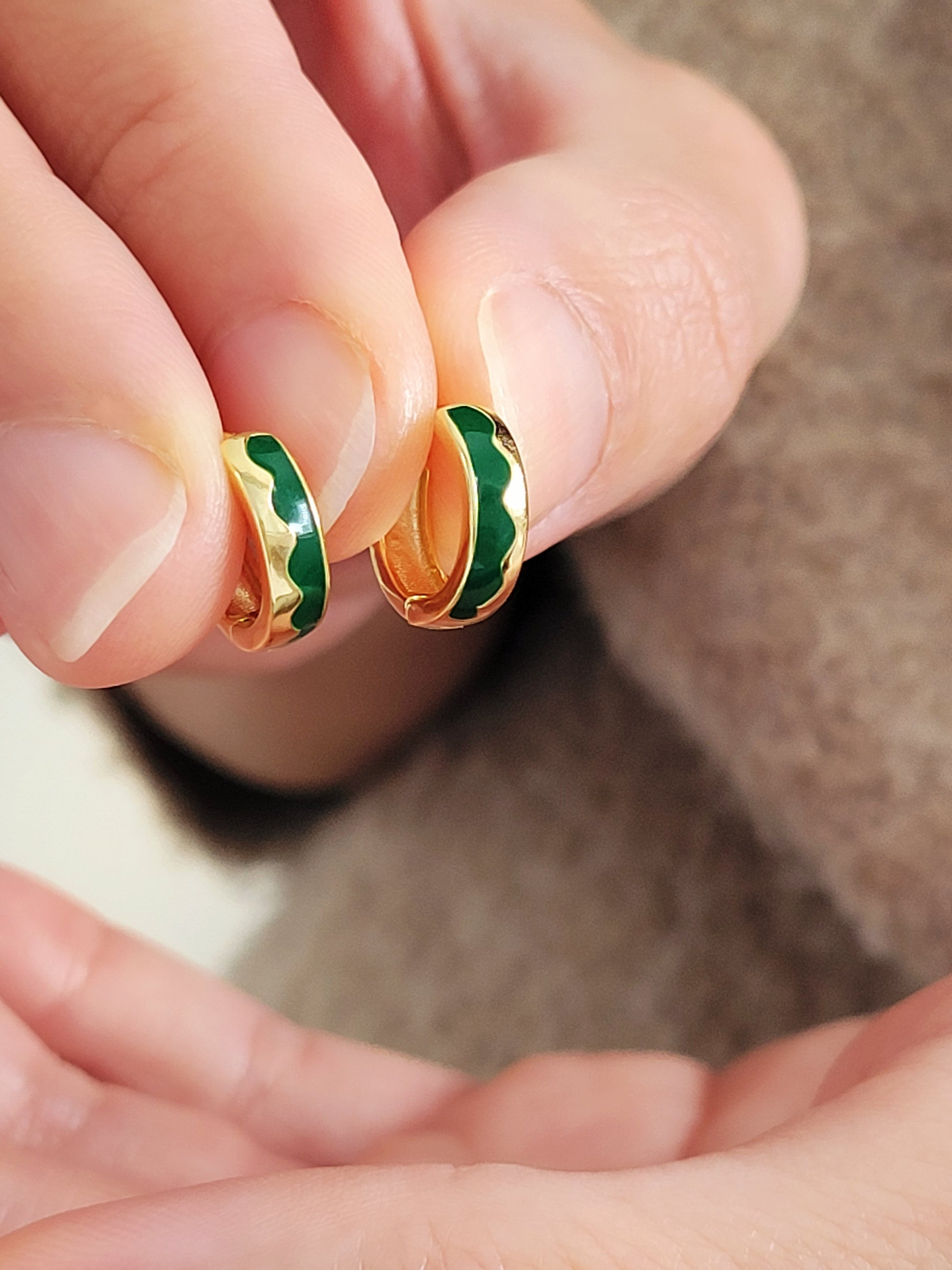 Gold and green earrings held between fingers against a neutral background