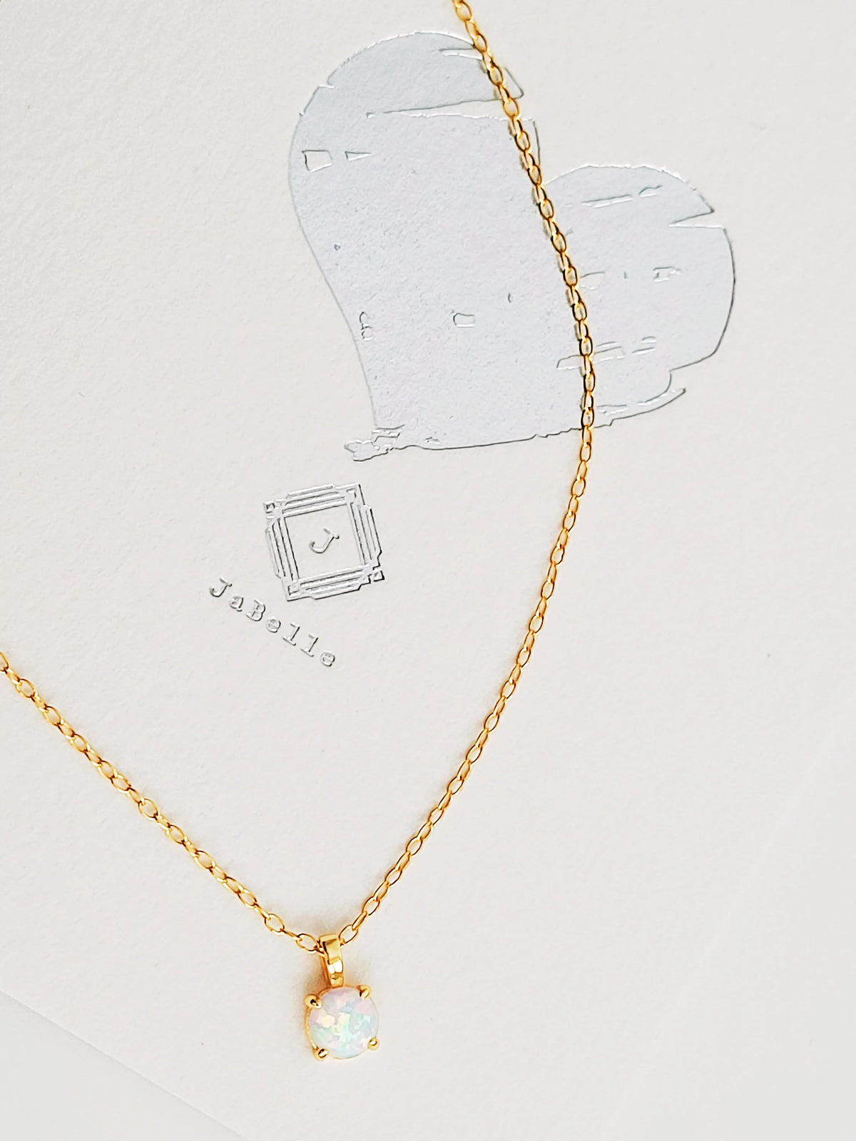 A delicate gold chain necklace with a small opal pendant holding a round, milky white opal gemstone, displayed on a textured white card with a silver heart illustration.