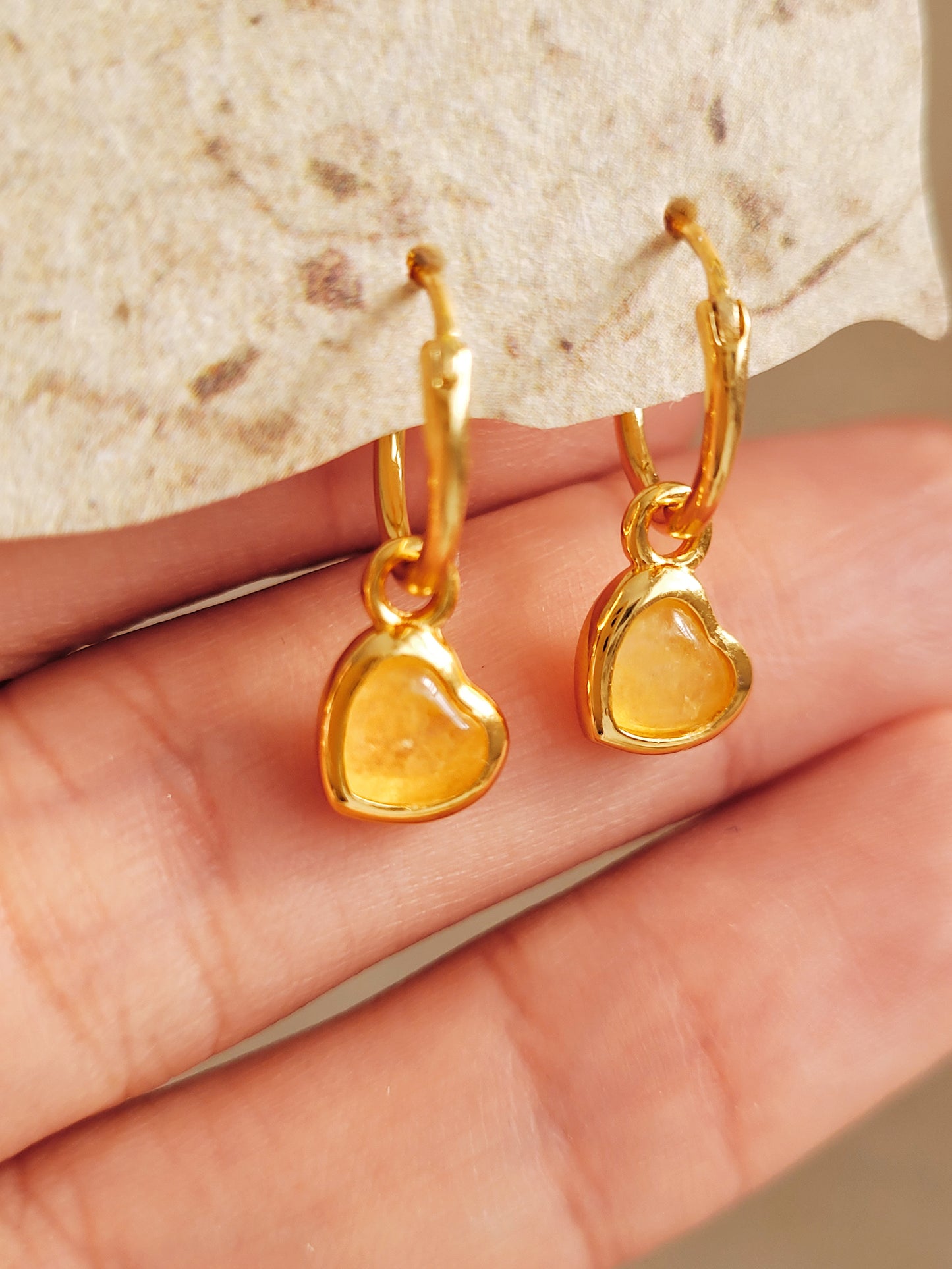 Gold heart crystal earrings held by a hand against a stone background