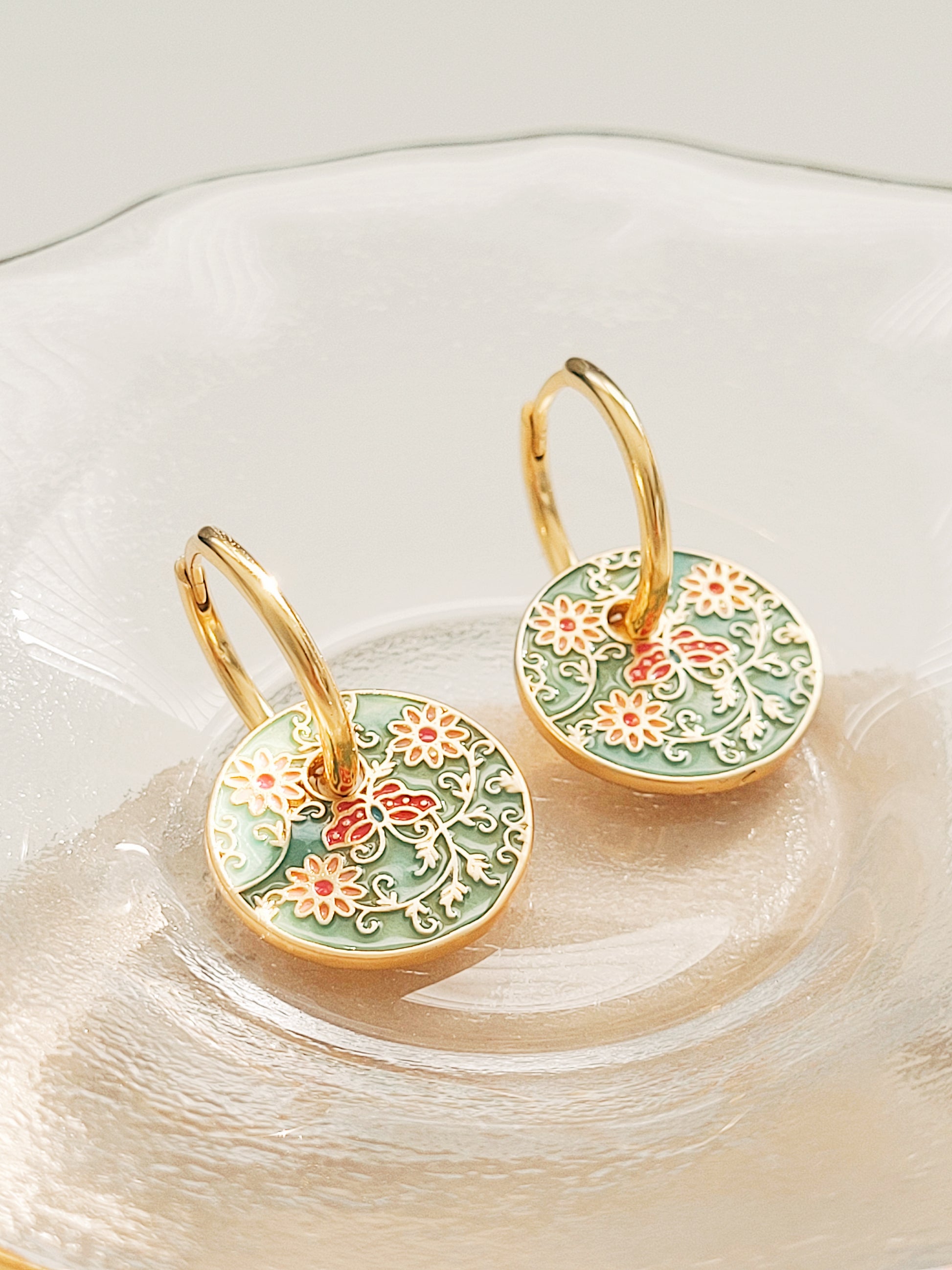 Gold enamel disc earrings with an Art Nouveau floral design in green, coral, and gold. Featuring intricate floral details with delicate vines and flower patterns, displayed on a clear glass dish, highlighting their vintage-inspired craftsmanship.