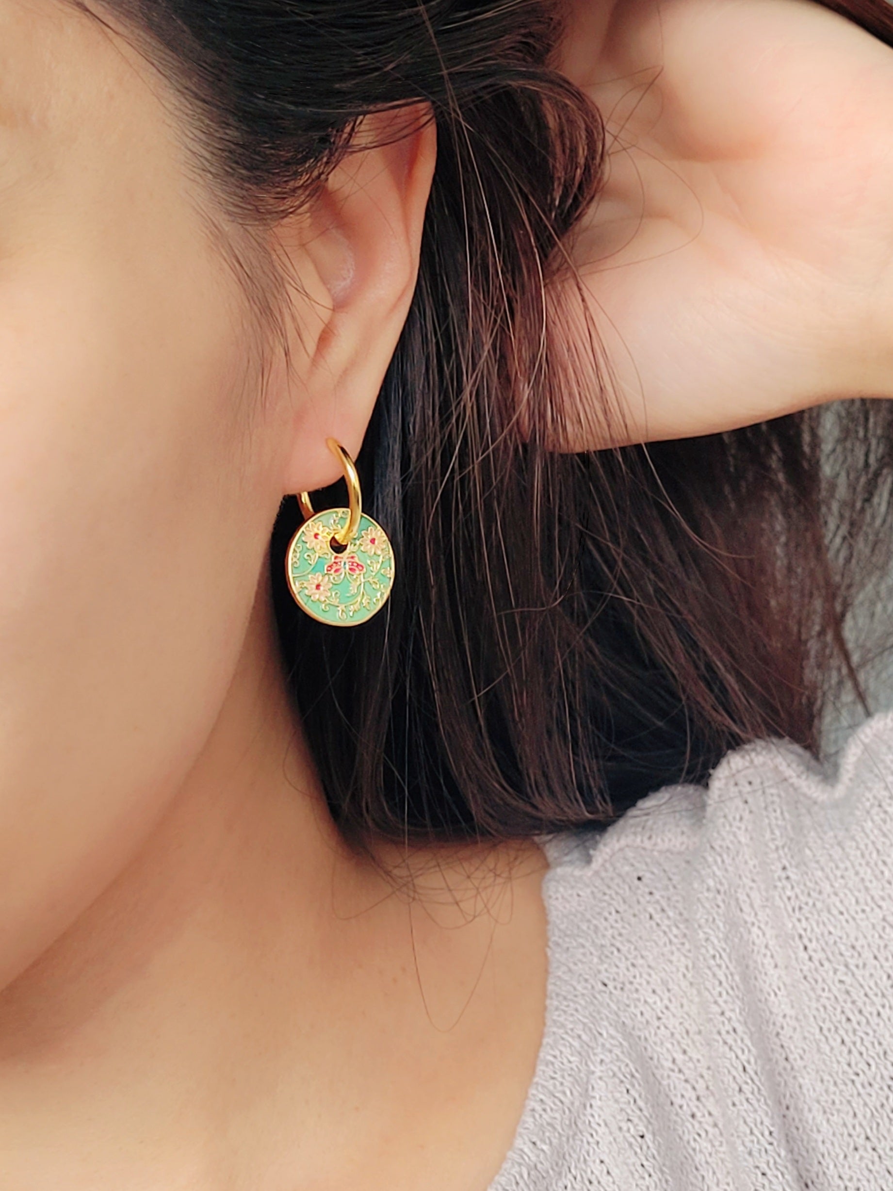 Gold enamel disc earrings with an Art Nouveau floral design in green, coral, and gold. Featuring intricate floral details with delicate vines and flower patterns, displayed on a clear glass dish, highlighting their vintage-inspired craftsmanship.