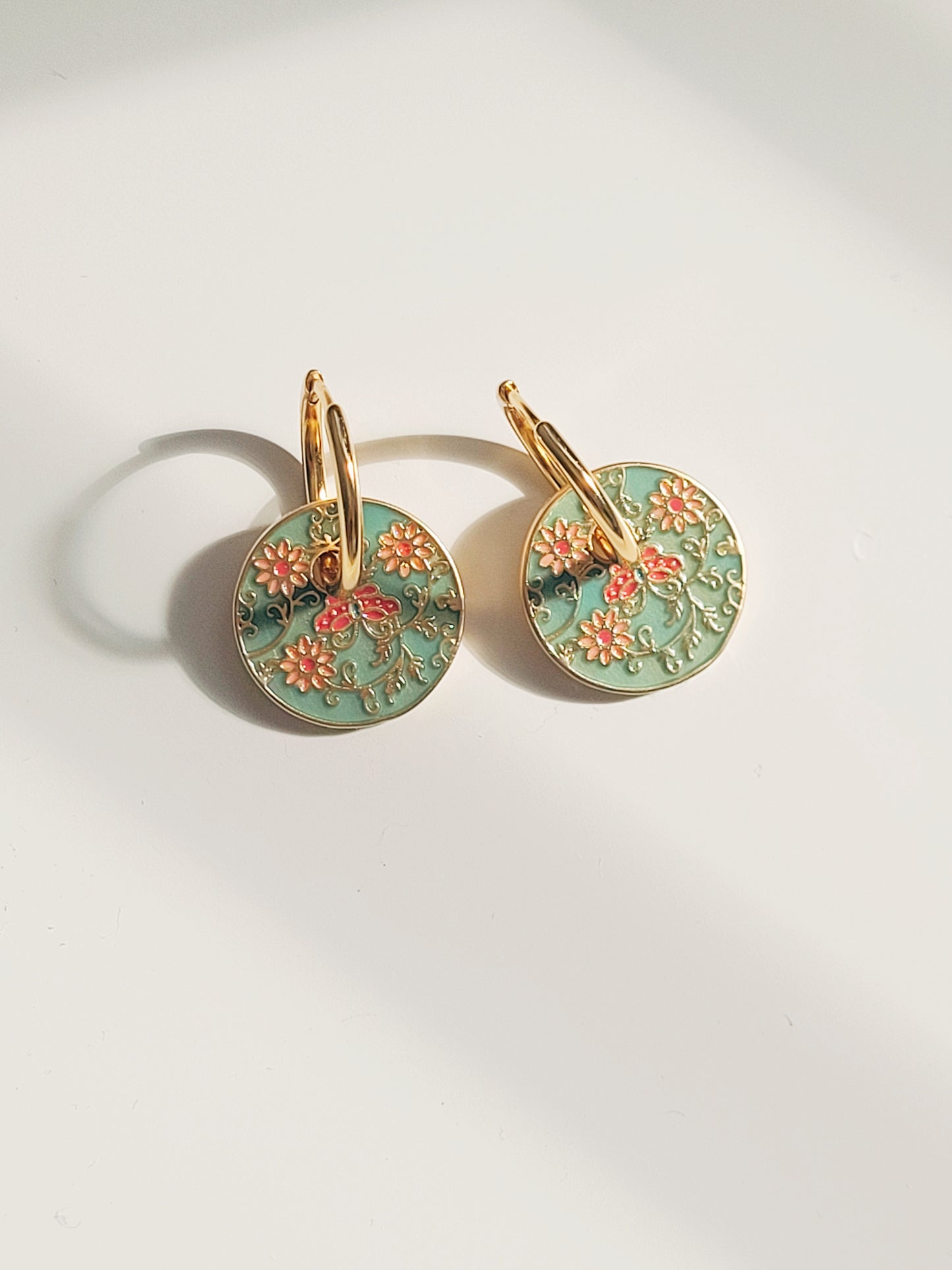 Gold enamel disc earrings with an Art Nouveau floral design in green, coral, and gold. Featuring intricate floral details with delicate vines and flower patterns, displayed on a clear glass dish, highlighting their vintage-inspired craftsmanship.