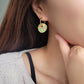 Gold enamel disc earrings with an Art Nouveau floral design in green, coral, and gold. Featuring intricate floral details with delicate vines and flower patterns, displayed on a clear glass dish, highlighting their vintage-inspired craftsmanship.