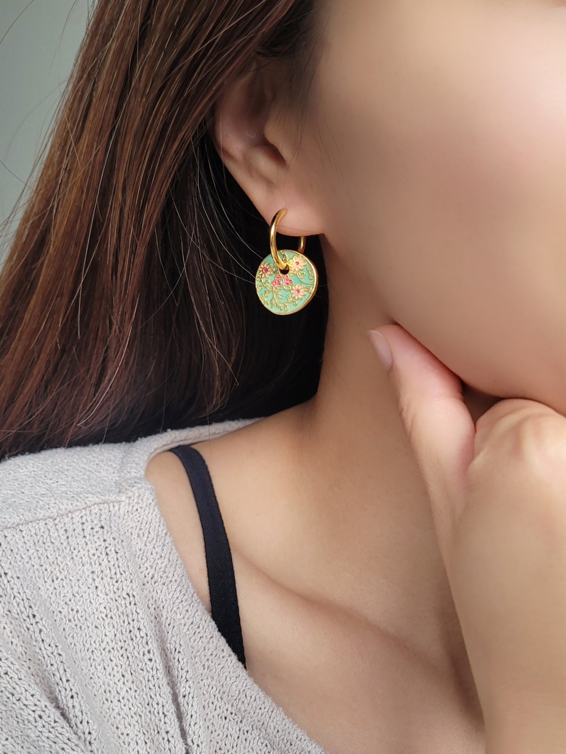 Gold enamel disc earrings with an Art Nouveau floral design in green, coral, and gold. Featuring intricate floral details with delicate vines and flower patterns, displayed on a clear glass dish, highlighting their vintage-inspired craftsmanship.