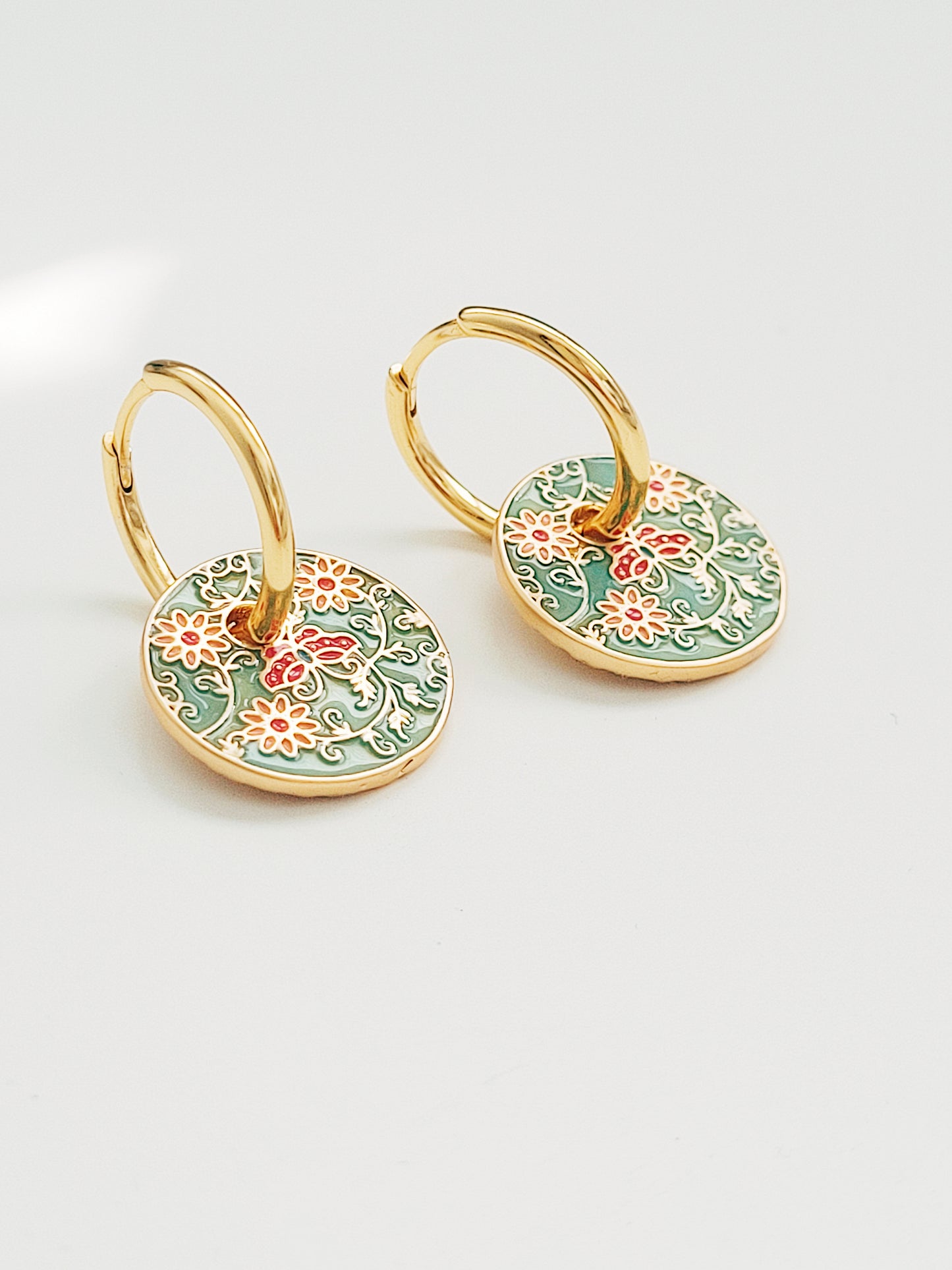 Gold enamel disc earrings with an Art Nouveau floral design in green, coral, and gold. Featuring intricate floral details with delicate vines and flower patterns, displayed on a clear glass dish, highlighting their vintage-inspired craftsmanship.