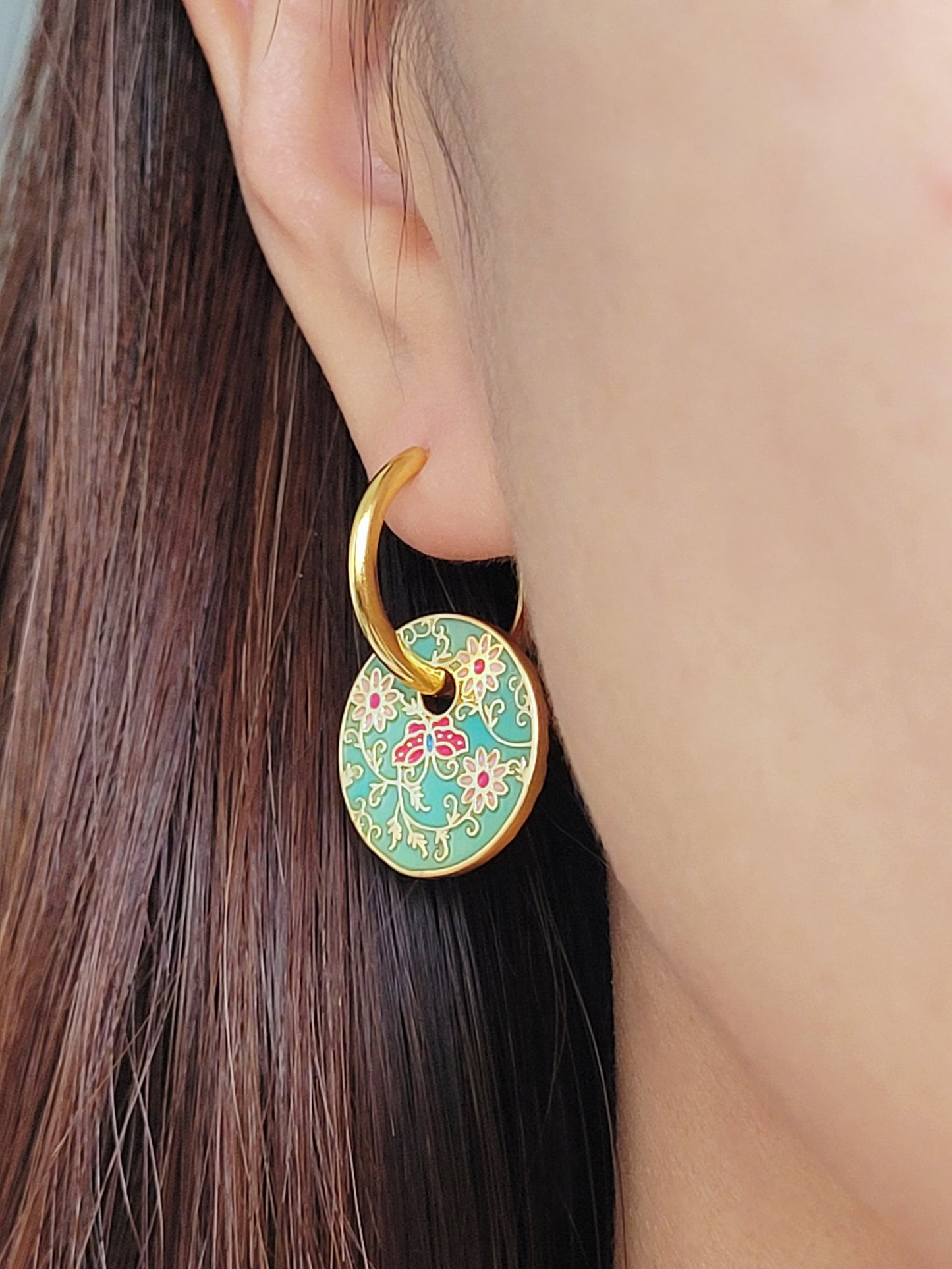 Gold enamel disc earrings with an Art Nouveau floral design in green, coral, and gold. Featuring intricate floral details with delicate vines and flower patterns, displayed on a clear glass dish, highlighting their vintage-inspired craftsmanship.