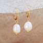 Pair of gold-plated huggie hoop earrings with freshwater pearl drops displayed on a textured fabric background.