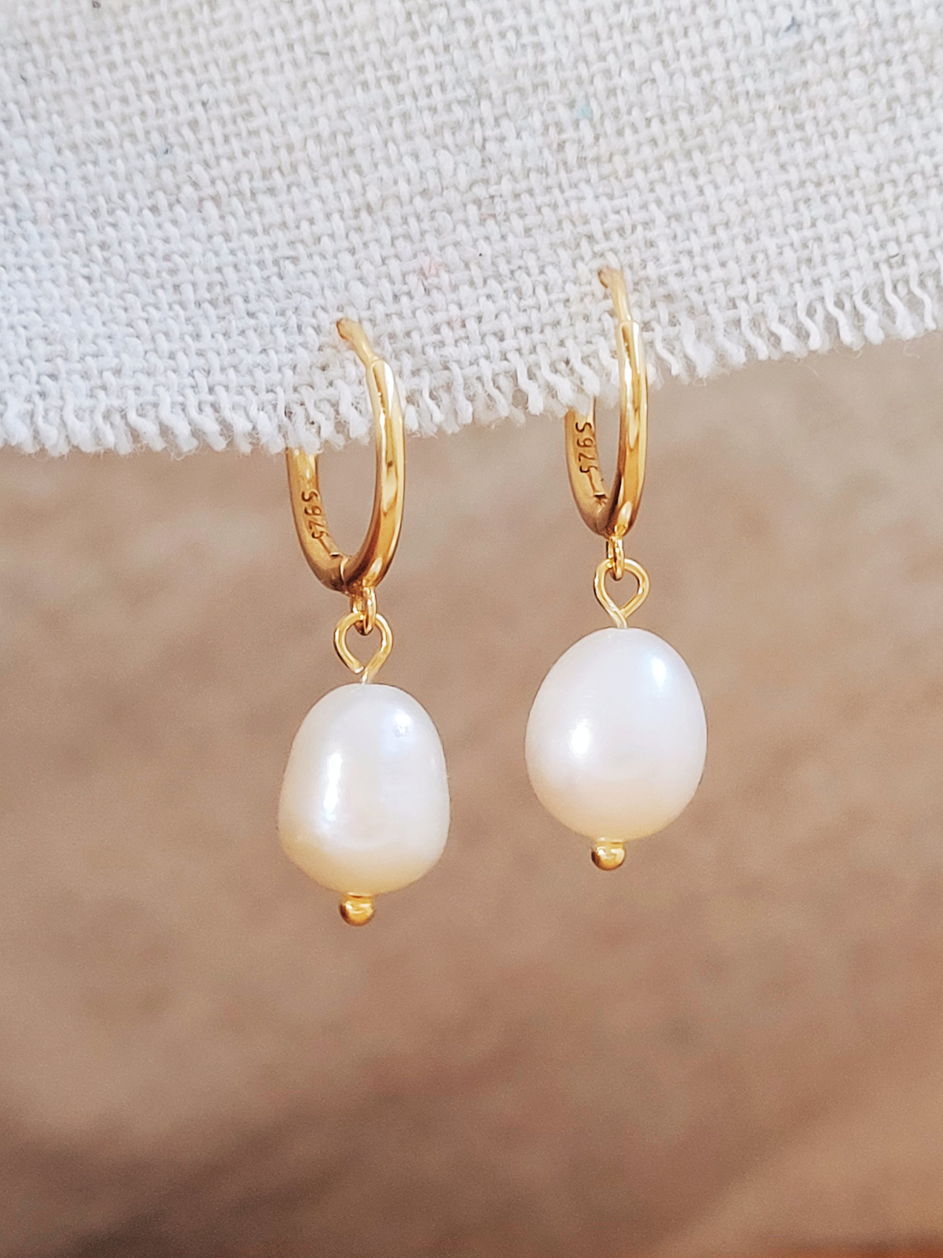 Pair of gold-plated huggie hoop earrings with freshwater pearl drops displayed on a textured fabric background.