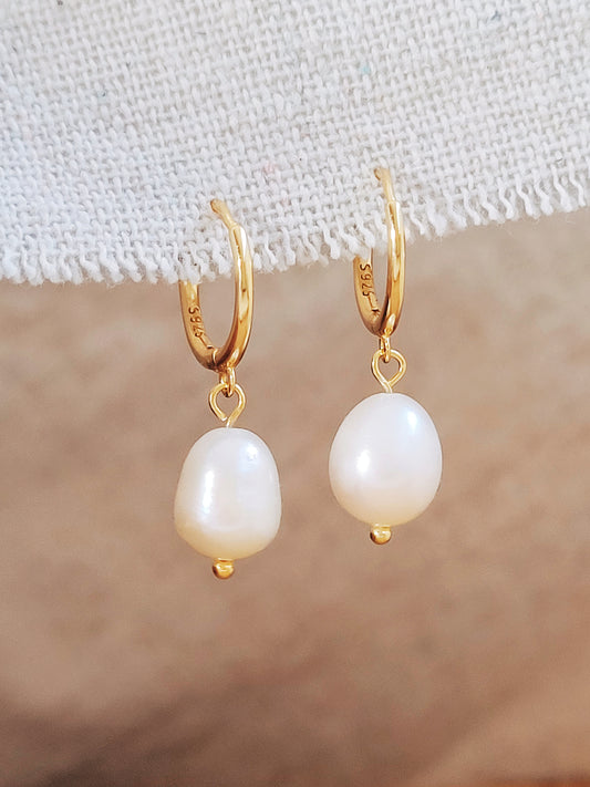 Pair of gold-plated huggie hoop earrings with freshwater pearl drops displayed on a textured fabric background.