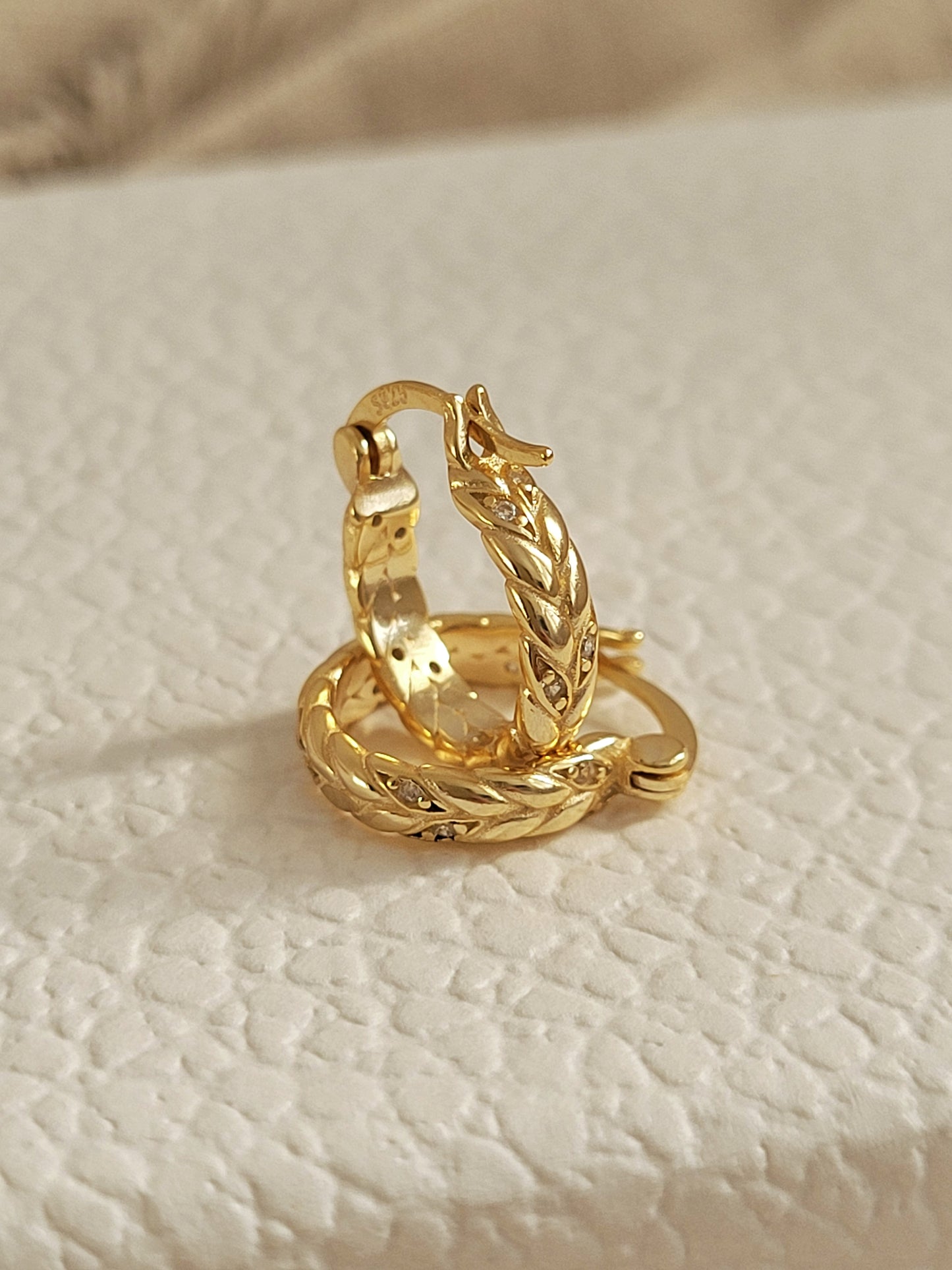 Gold plated braided hoop earring on a textured beige surface.