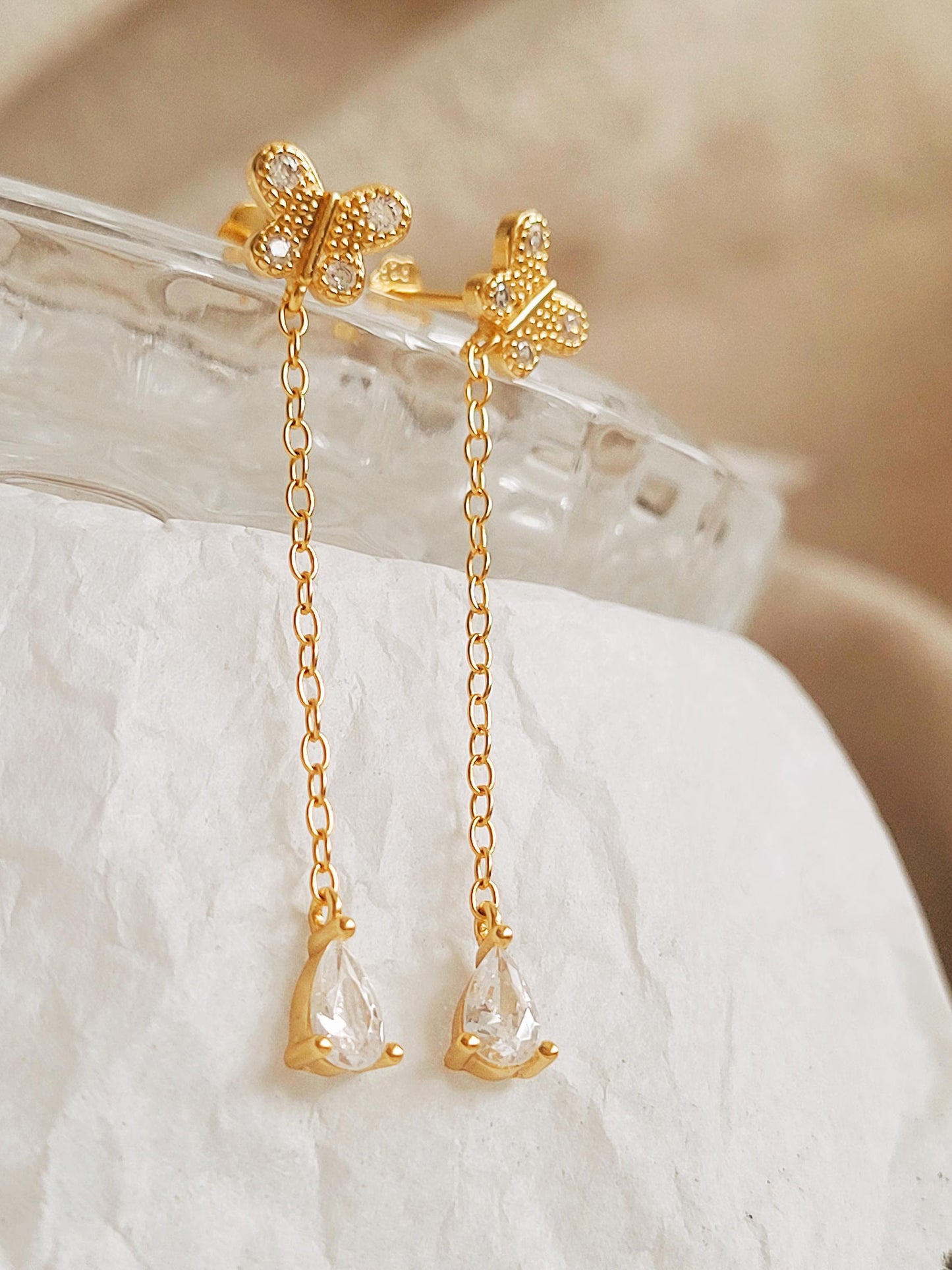 A pair of dainty gold butterfly earrings with chain drops on a white fabric background.