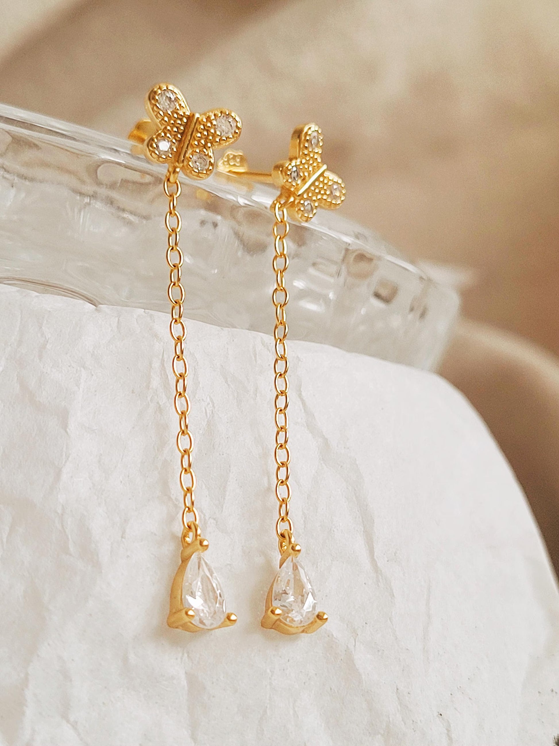 A pair of dainty gold butterfly earrings with chain drops on a white fabric background.
