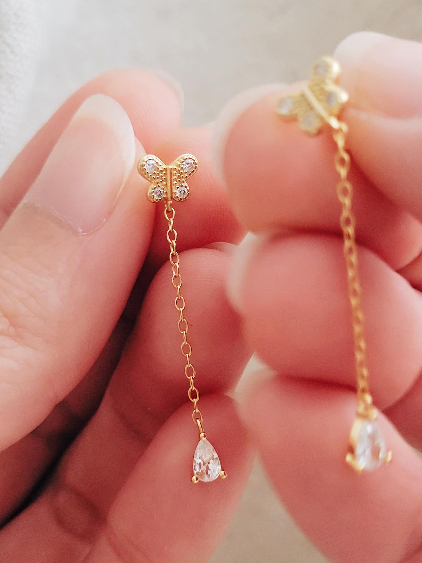 Gold butterfly and teardrop earrings held between fingers against a neutral background