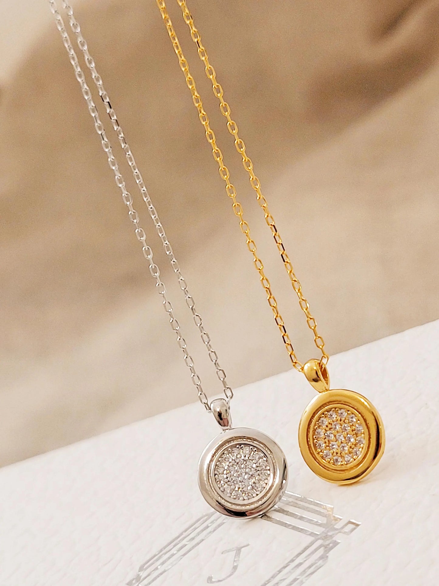 Two necklaces, one gold and one silver, with circular pendants on a beige fabric background.