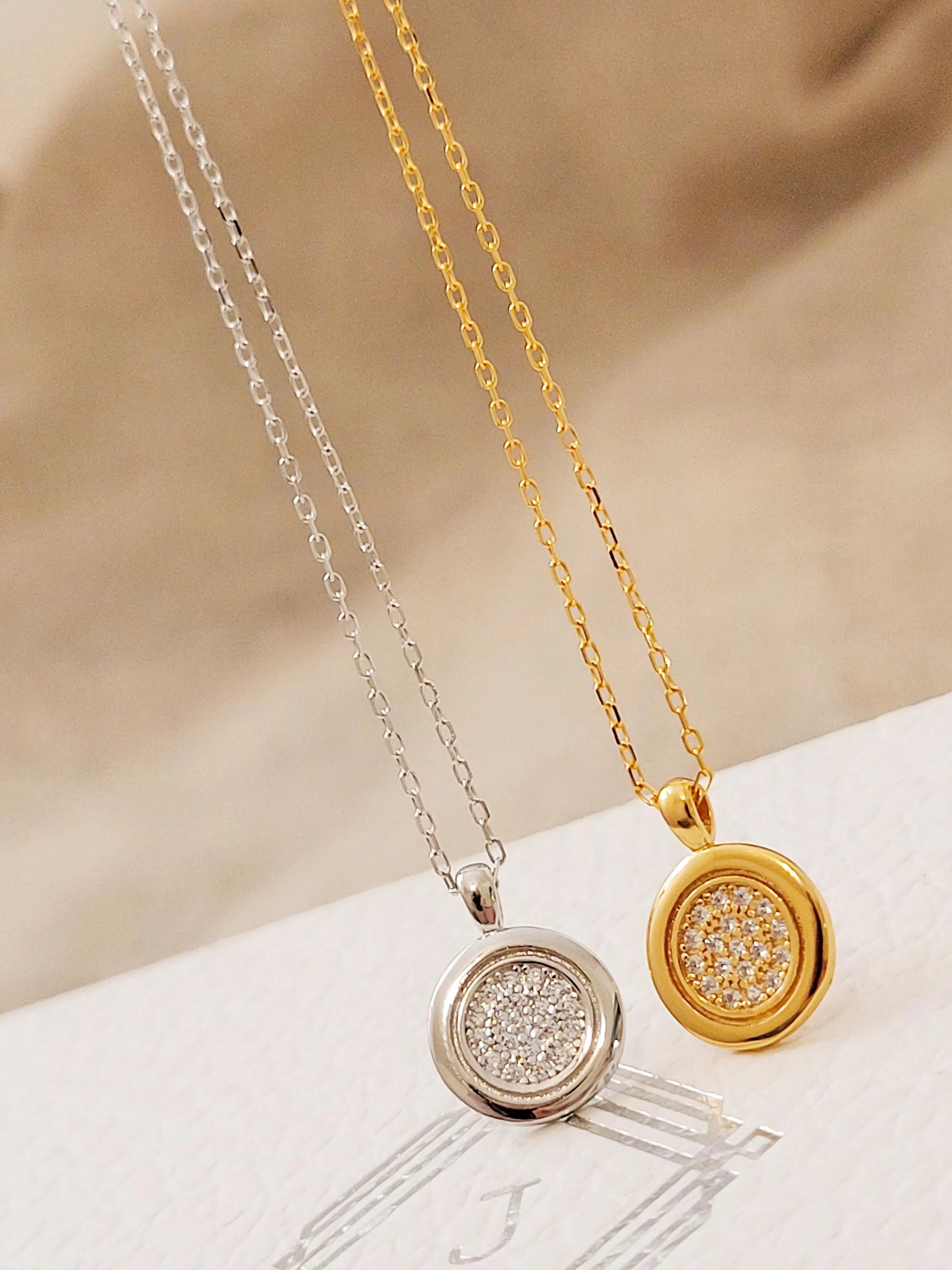 Two necklaces, one gold and one silver, with circular pendants on a beige fabric background.