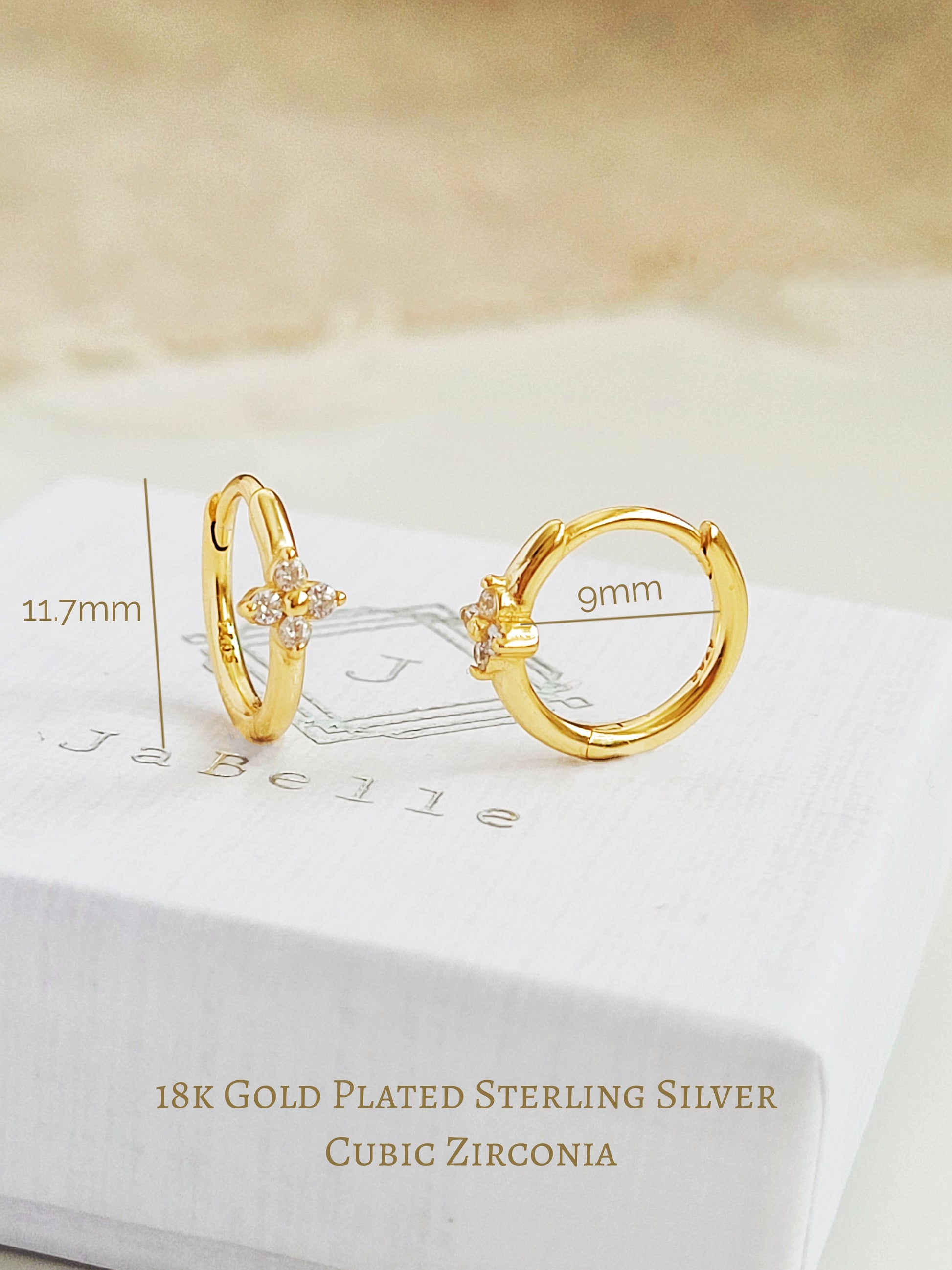 Close up of a pair of gold clover huggie earrings displayed on a jewellery box with dimensions.