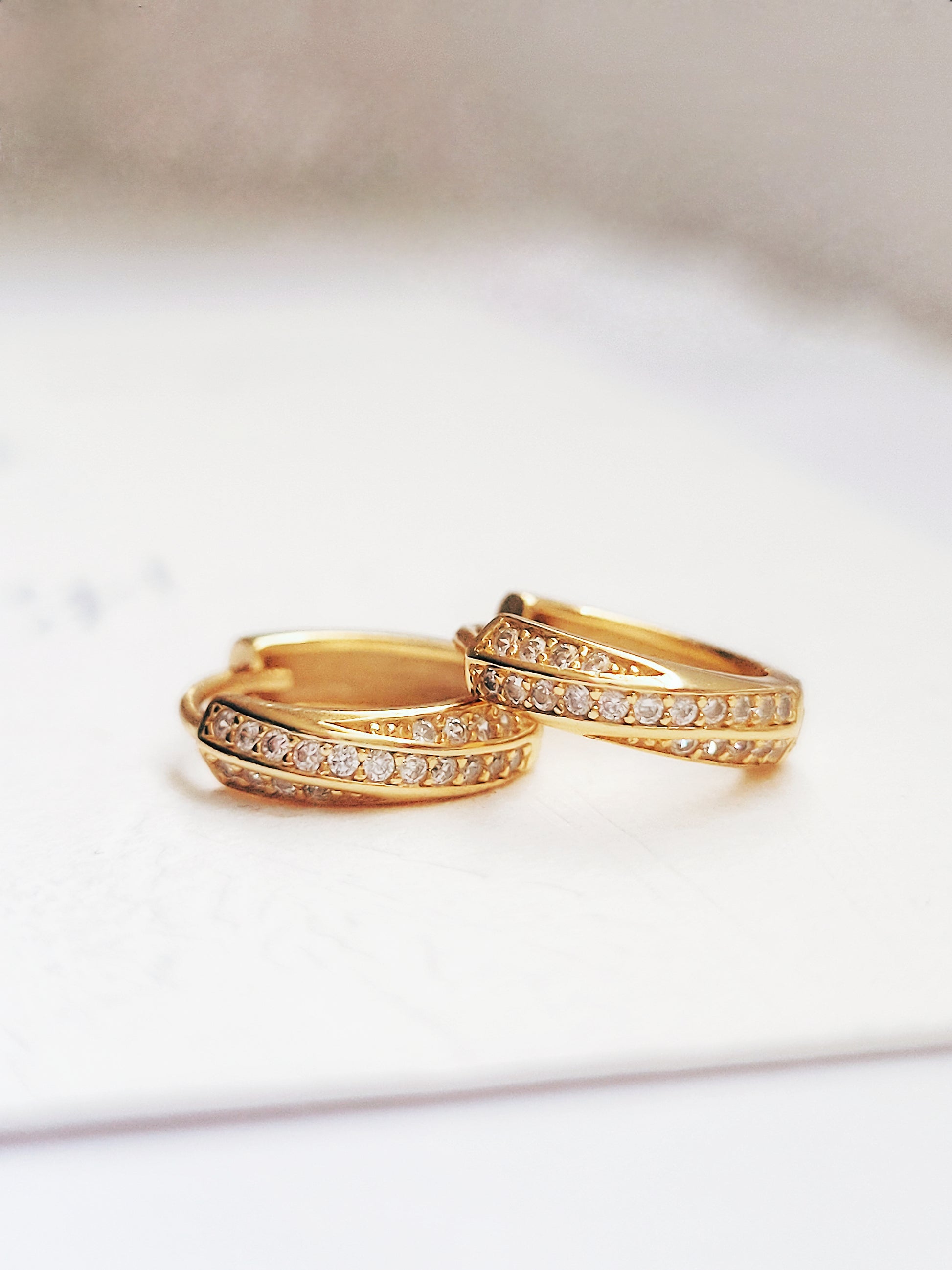 A pair of gold crisscross huggie hoop earrings. The earrings are adorned with small Zirconia stones, set along two intersecting bands, giving them a sparkling, elegant look.