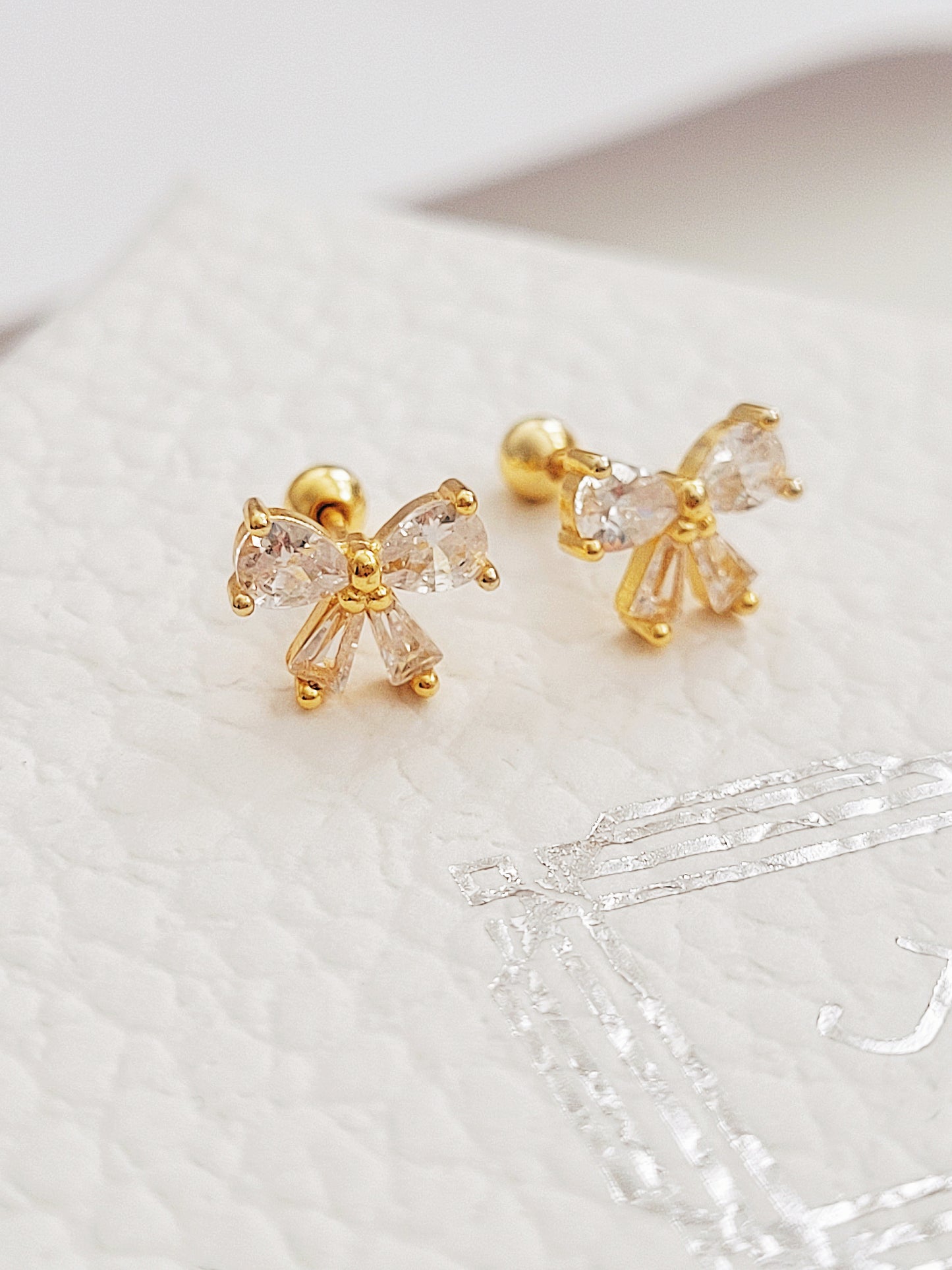 Close up of a woman&#39;s ear wearing dainty bow stud earrings with sparkling cubic zirconia and gold plating.