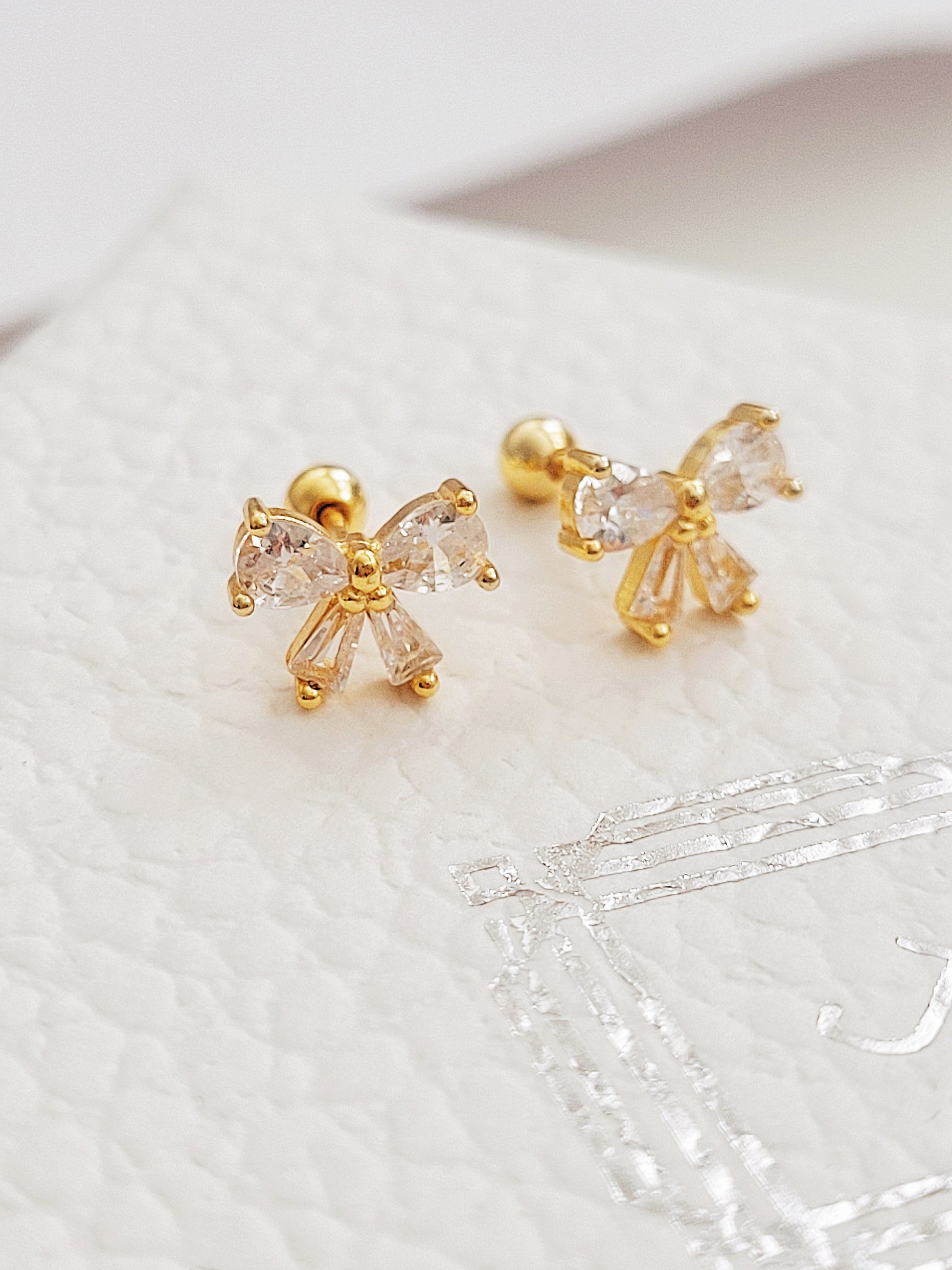 Close up of a woman&#39;s ear wearing dainty bow stud earrings with sparkling cubic zirconia and gold plating.