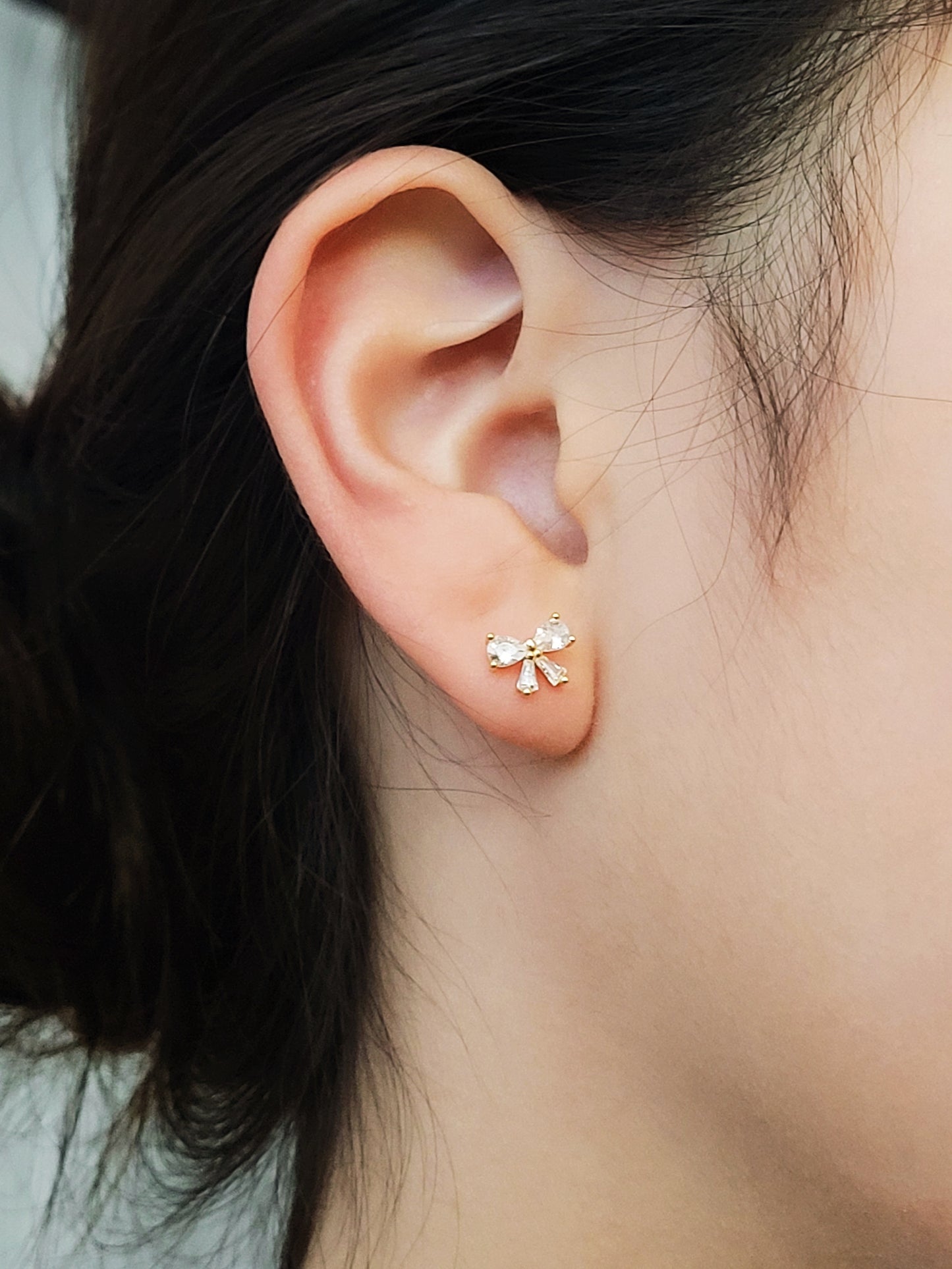 Close up of a woman&#39;s ear wearing dainty bow stud earrings with sparkling cubic zirconia and gold plating.