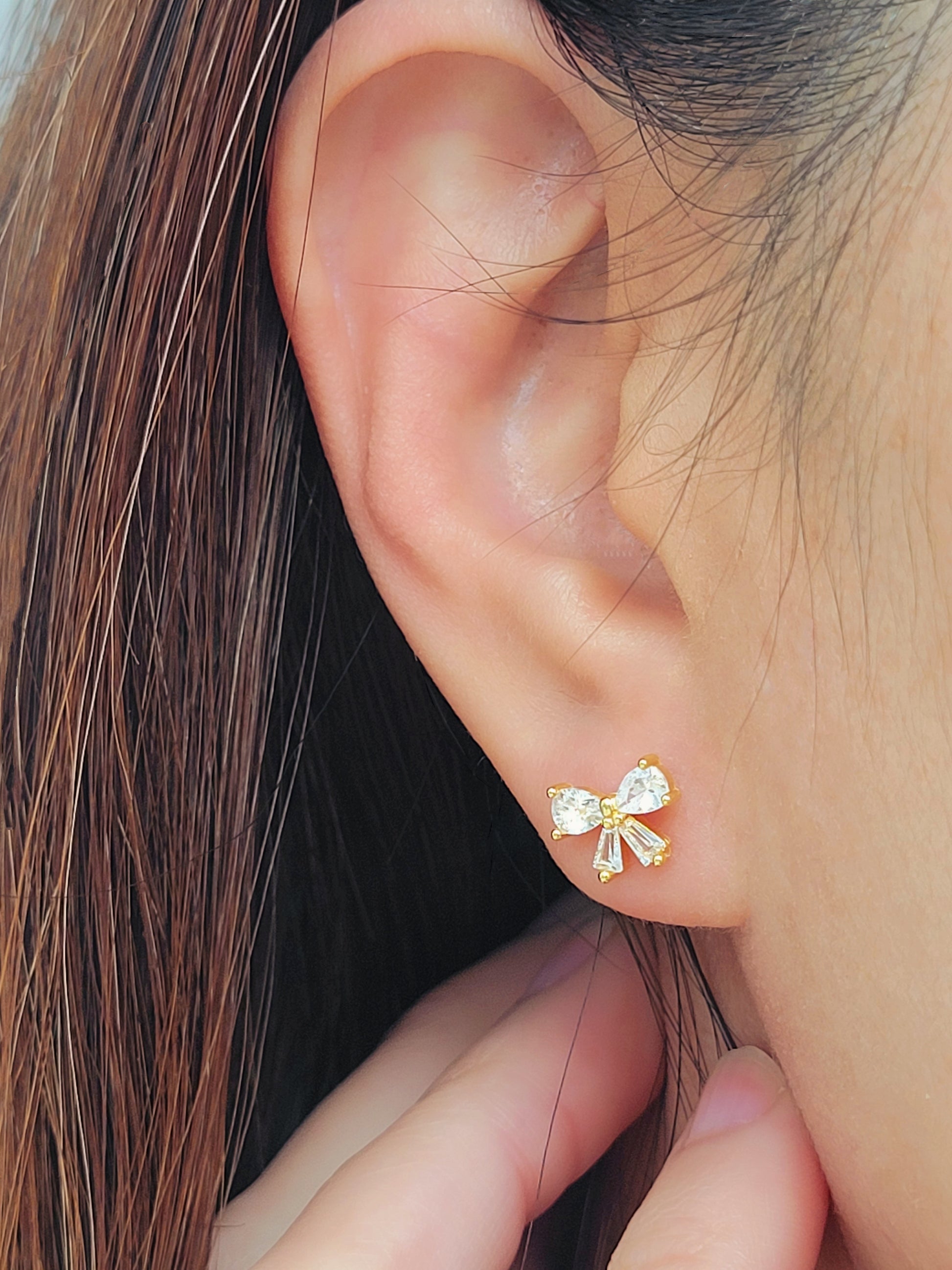 Close up of a woman&#39;s ear wearing dainty bow stud earrings with sparkling cubic zirconia and gold plating.