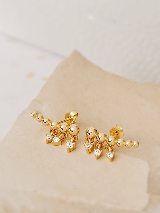 Minimalist gold climber earrings featuring dainty dangling accents, resting on a soft beige stone background.