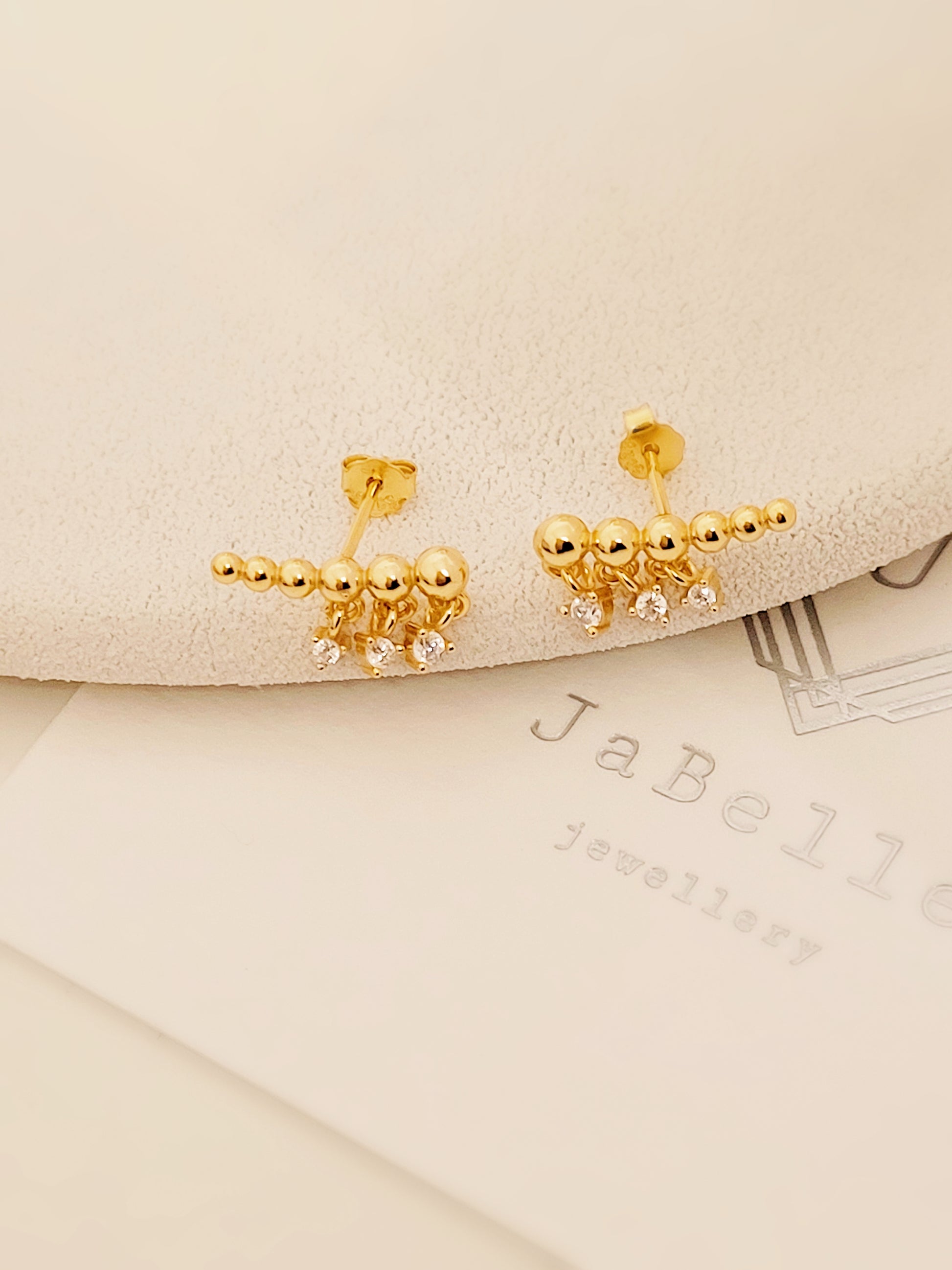 Minimalist gold climber earrings featuring dainty dangling accents, resting on a soft beige background.