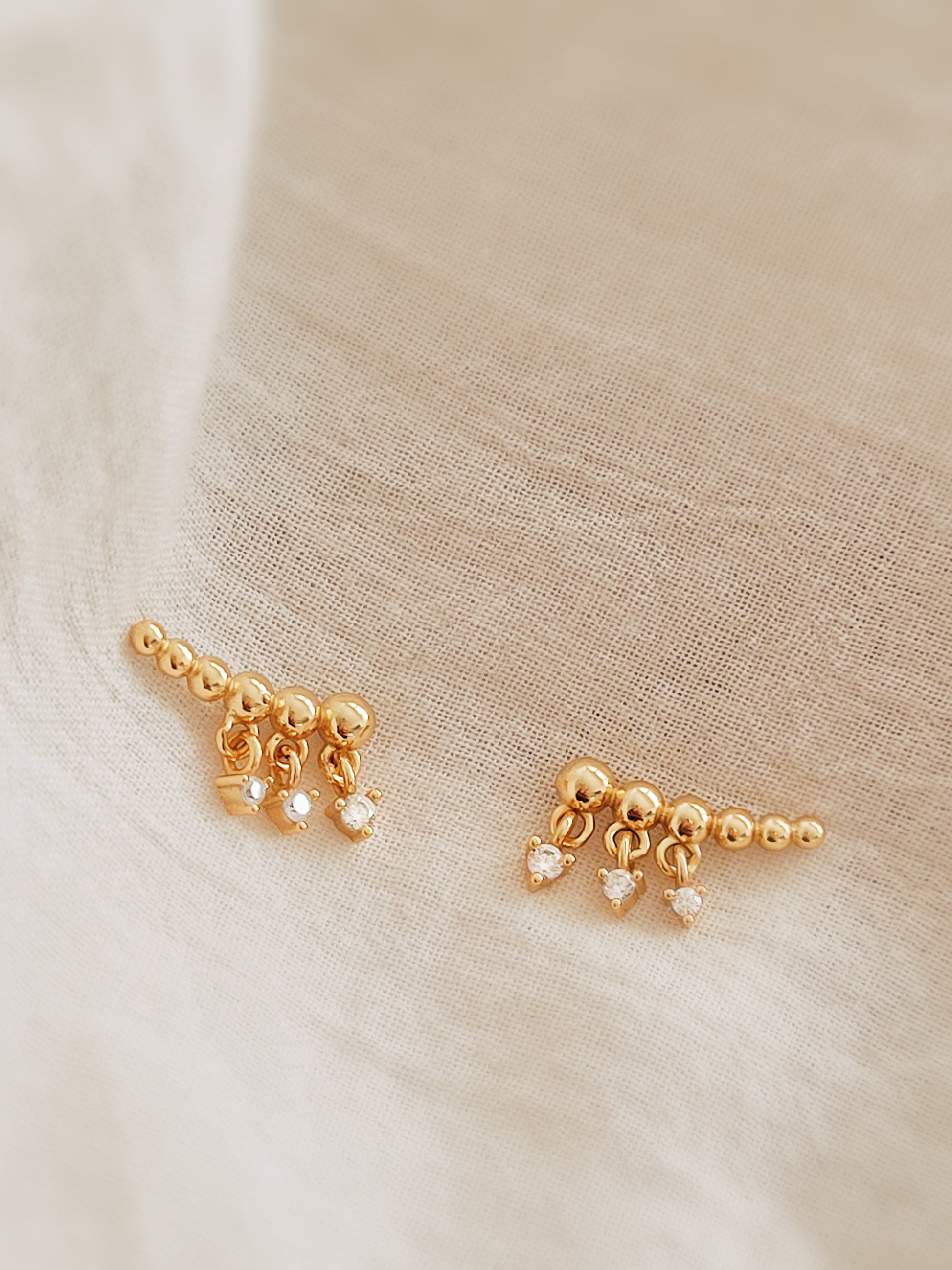 Minimalist gold climber earrings featuring dainty dangling accents, resting on a soft beige background.