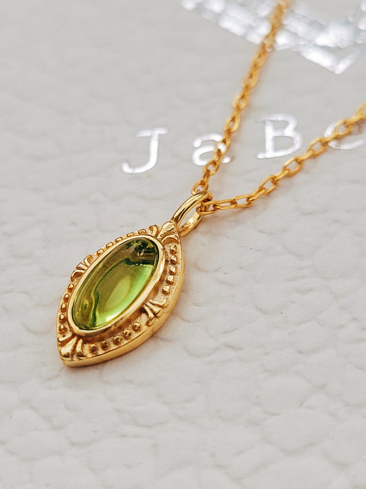 Close-up of a vintage-inspired gold pendant necklace featuring an oval green gemstone on a delicate gold chain, displayed on a textured background.