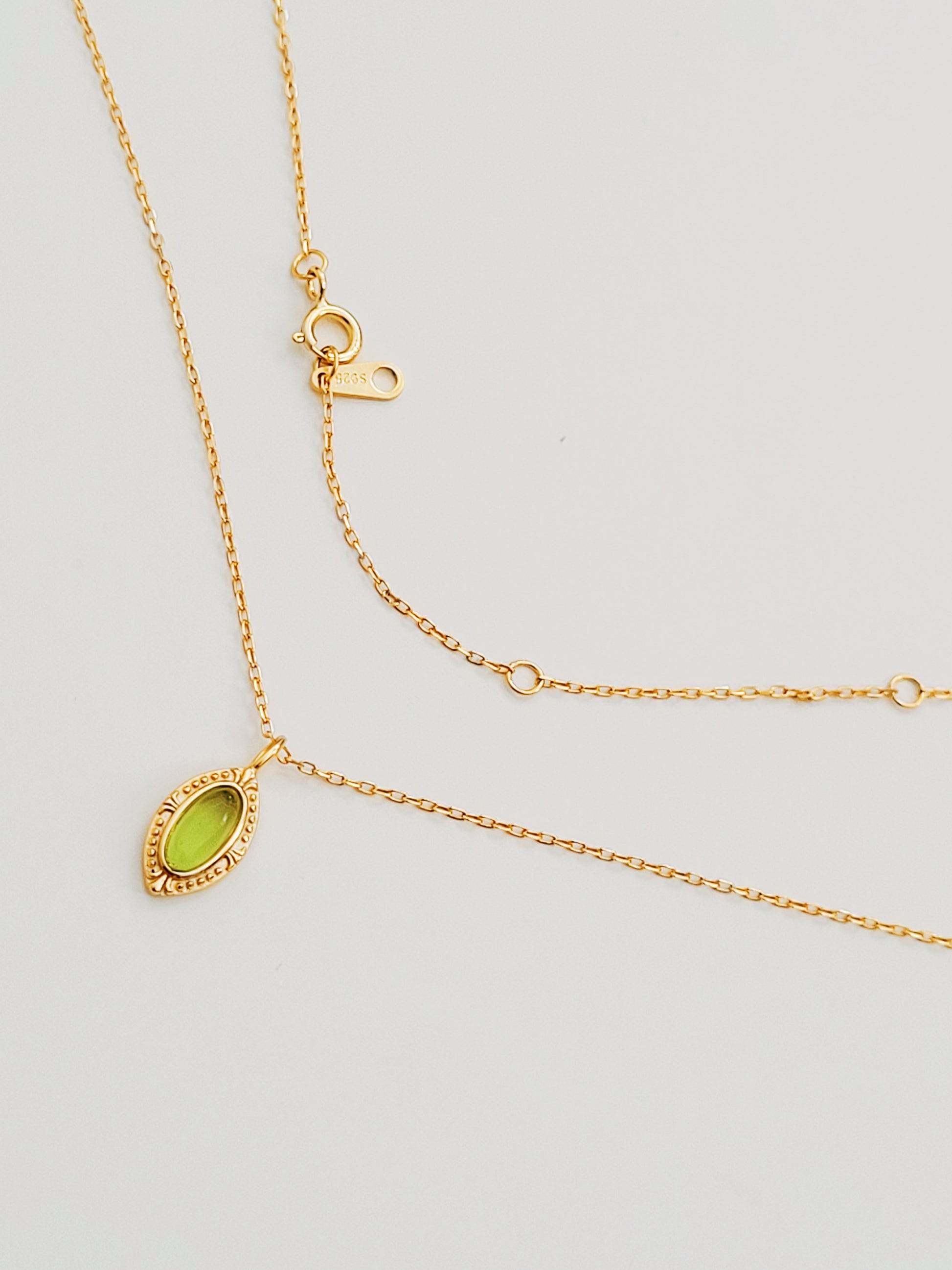 Dainty gold-tone pendant necklace with a sparkling oval green stone, lying on a cream surface with subtle branding in the background.