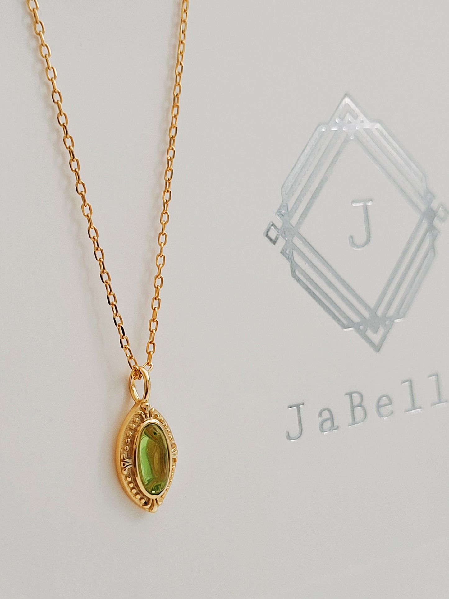 Close-up of a vintage-inspired gold pendant necklace featuring an oval green gemstone on a delicate gold chain, displayed on a textured background.