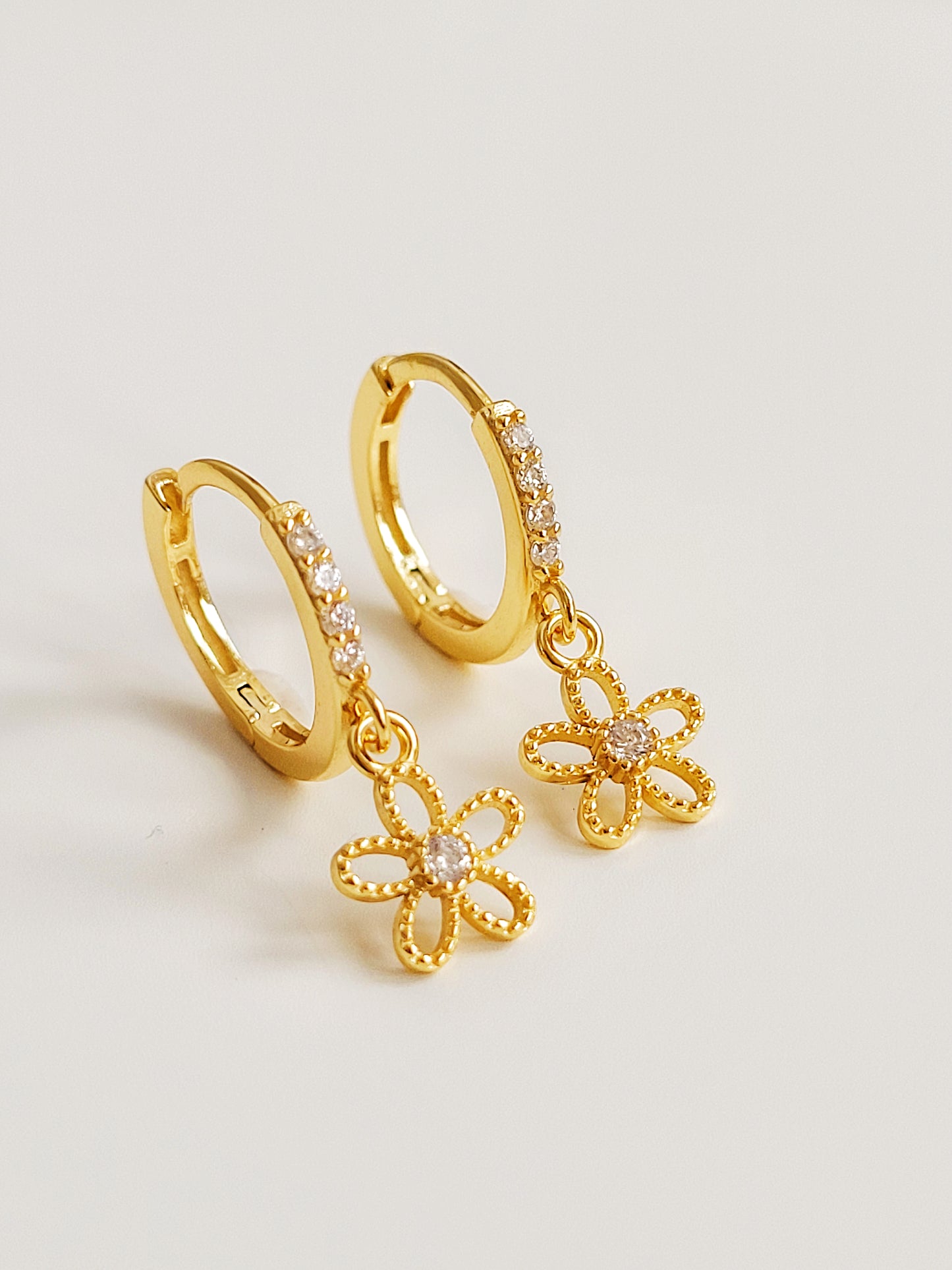 A pair of dainty gold huggie earrings with delicate flower charms on a white surface.