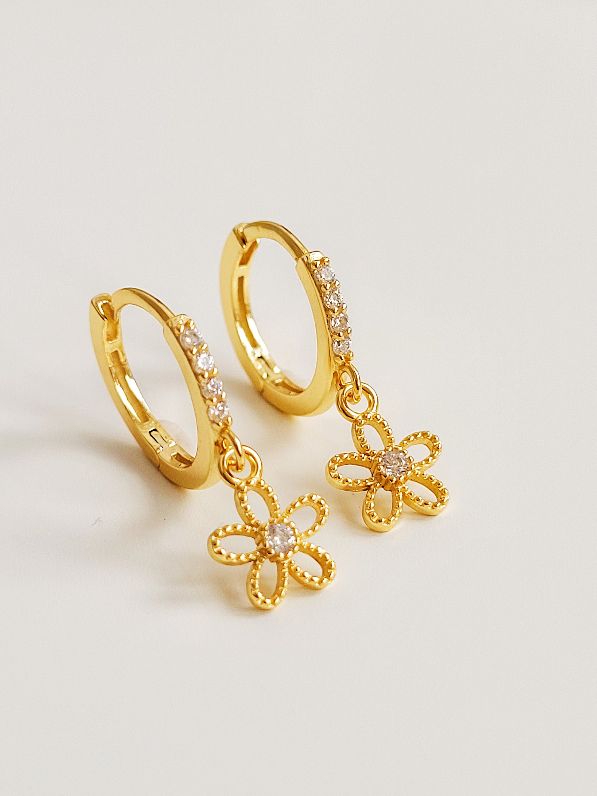 A pair of dainty gold huggie earrings with delicate flower charms on a white surface.