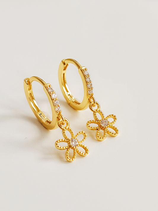 A pair of dainty gold huggie earrings with delicate flower charms on a white surface.