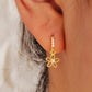 A close-up image of a woman's ear wearing a gold plated huggie earring with a small dangling flower design.