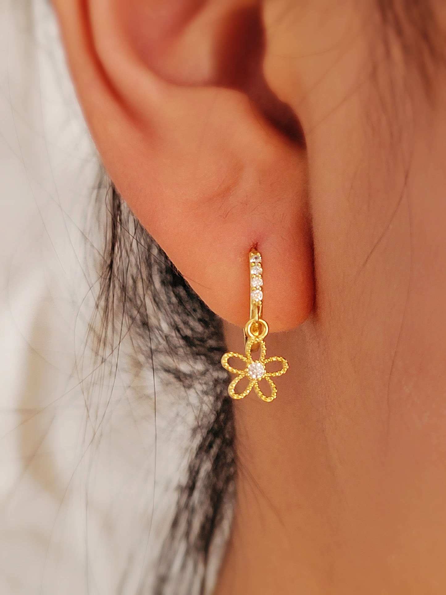A close-up image of a woman's ear wearing a gold plated huggie earring with a small dangling flower design.